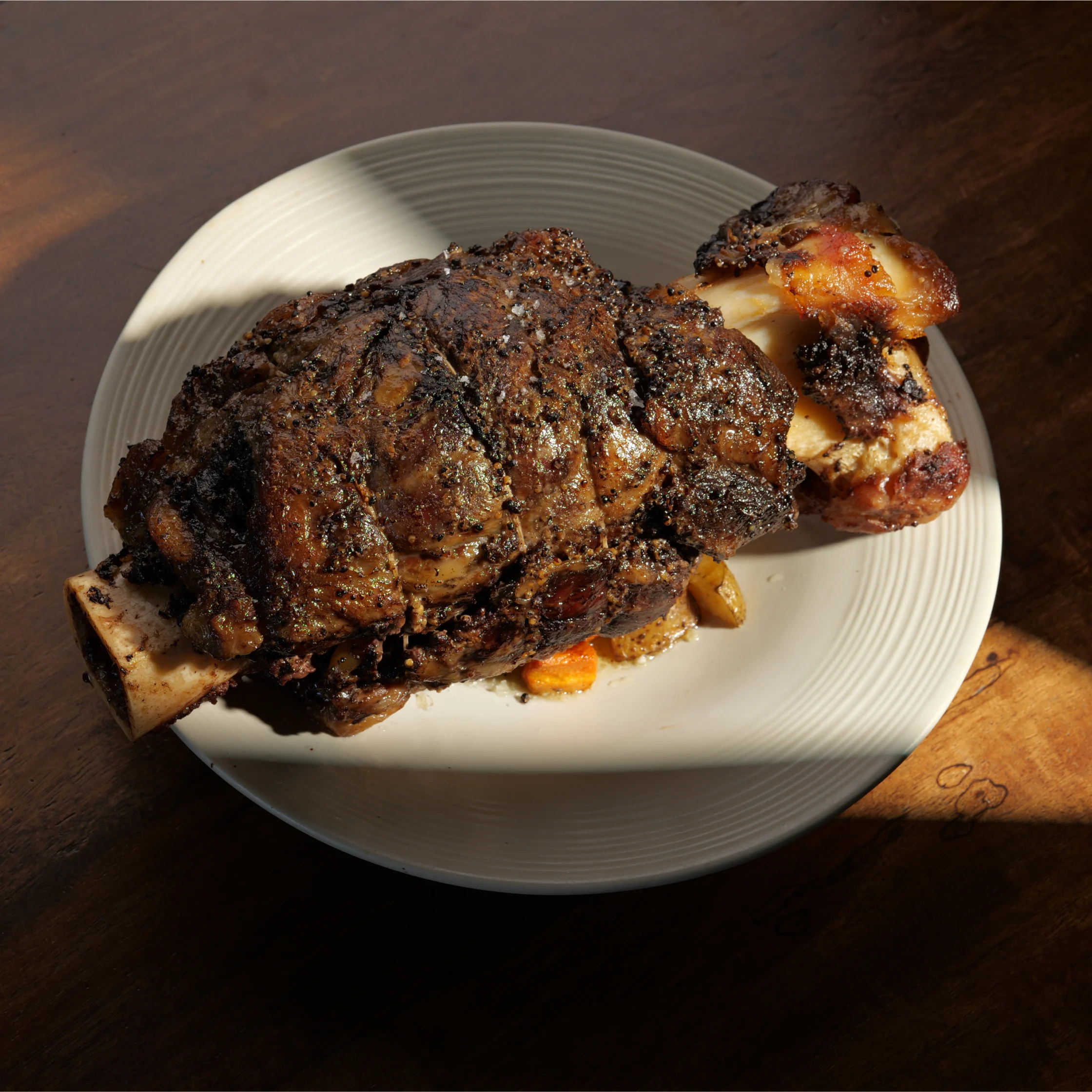 A large roasted beef shank served on a white plate, featuring deeply browned, pepper-crusted meat wrapped around a prominent marrow bone, with roasted vegetables tucked underneath.