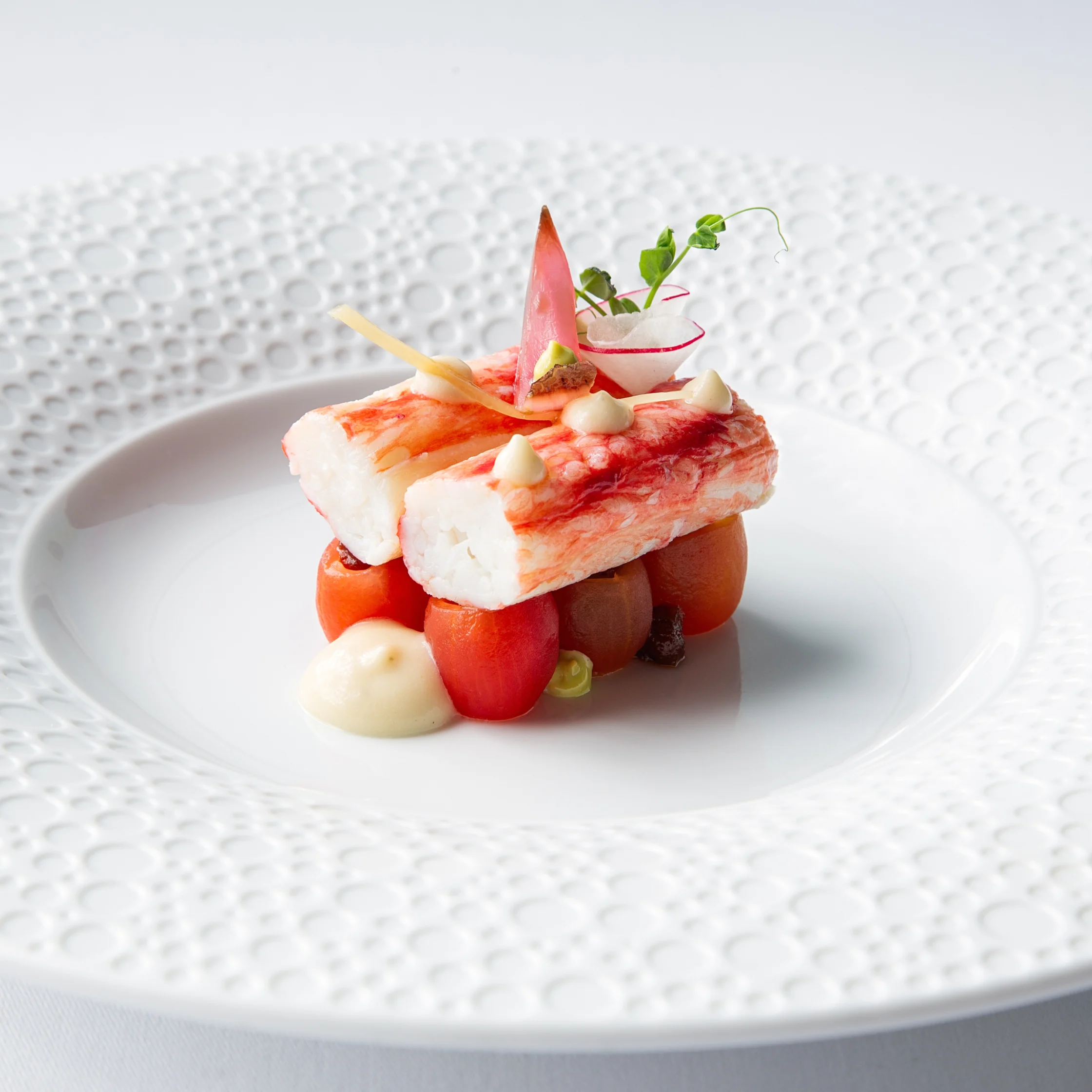 Signature BRONTË Alaskan king crab dish, artfully plated with crab meat over small tomatoes and finished with creamy dollops, delicate vegetable garnishes, and microgreens on a textured white plate.
