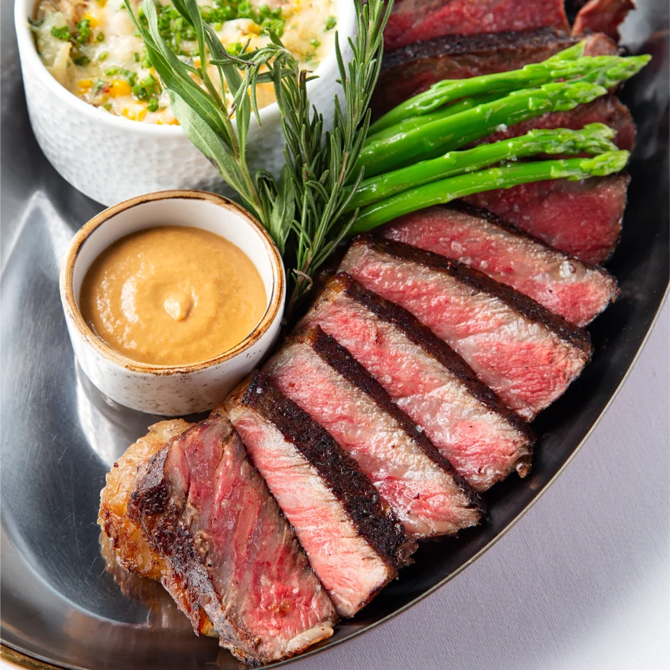 BRONTË bone-in New York strip steak sliced medium-rare and served with asparagus, rosemary, a creamy sauce, and a side of corn and brie gratin.