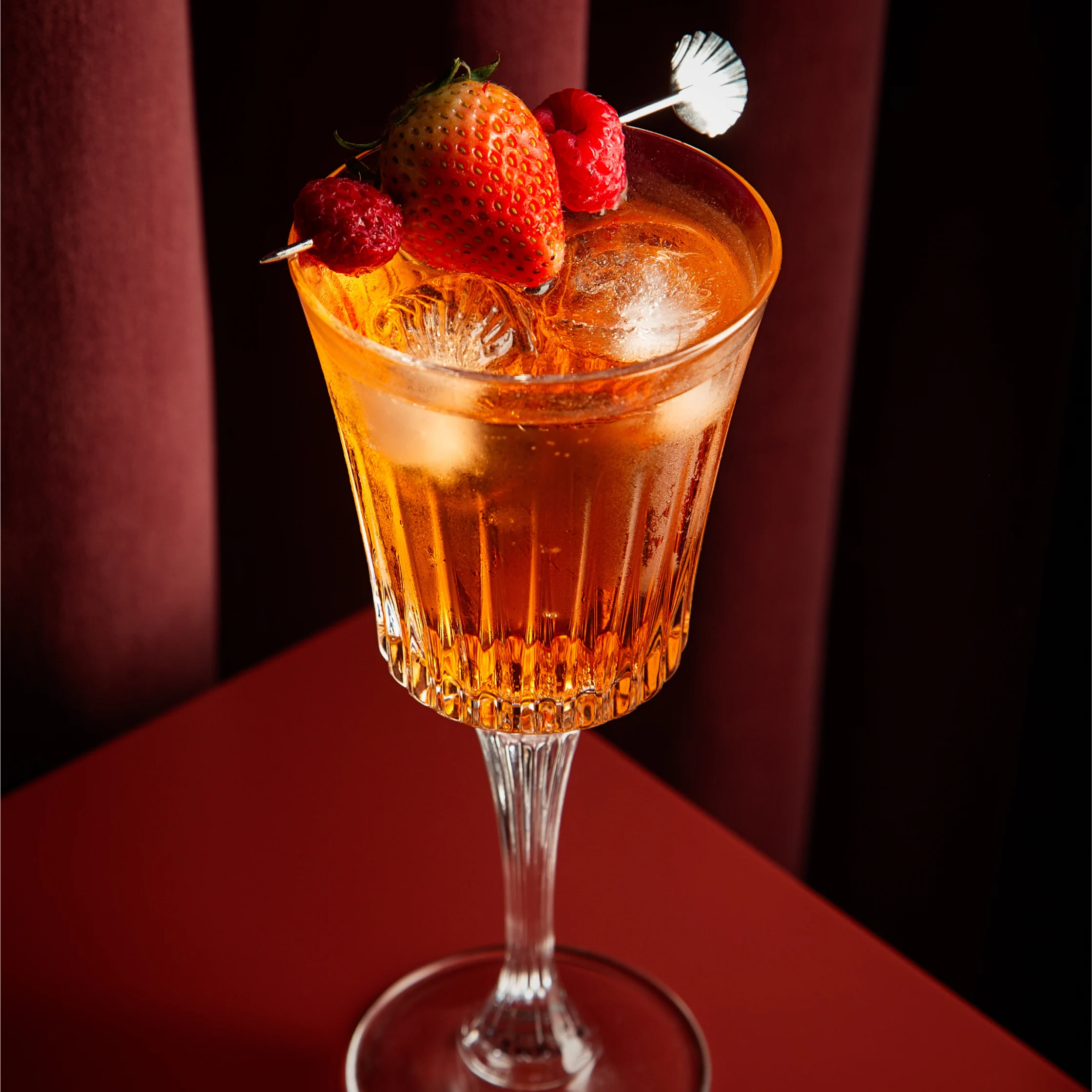 Signature BRONTË cocktail in a stemmed glass, garnished with a strawberry and raspberries, displayed on a red table against rich burgundy curtains.