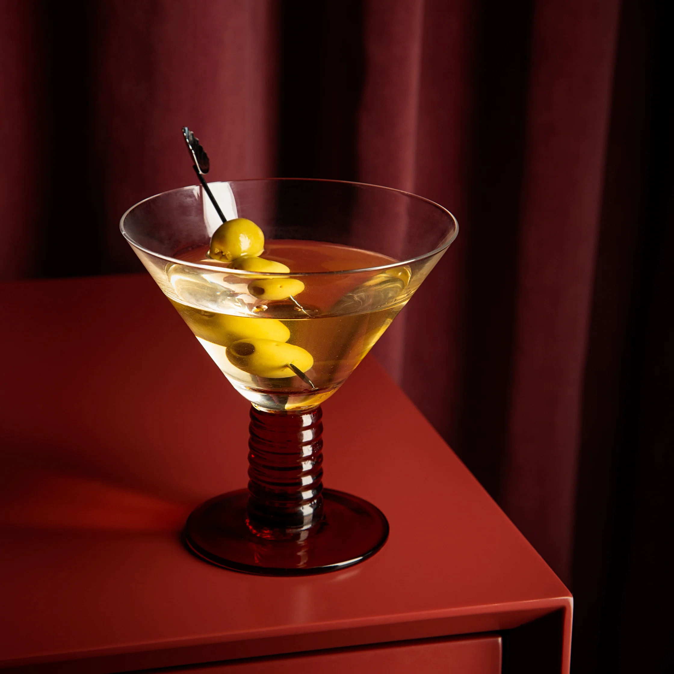 BRONTË dirty martini served in a wide cocktail glass with green olives on a skewer, presented on a red table in front of burgundy curtains.
