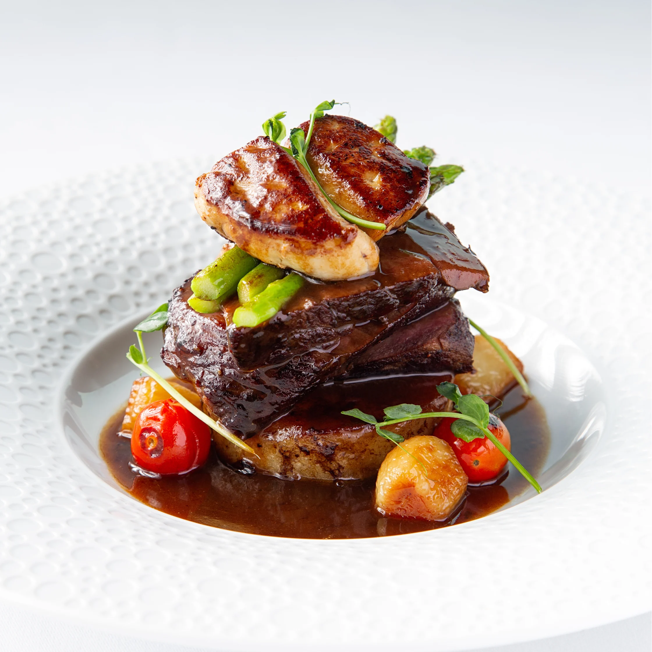 BRONTË Filet Rossini featuring a glazed beef fillet topped with seared foie gras and asparagus, served with roasted vegetables and rich jus on a white textured plate.