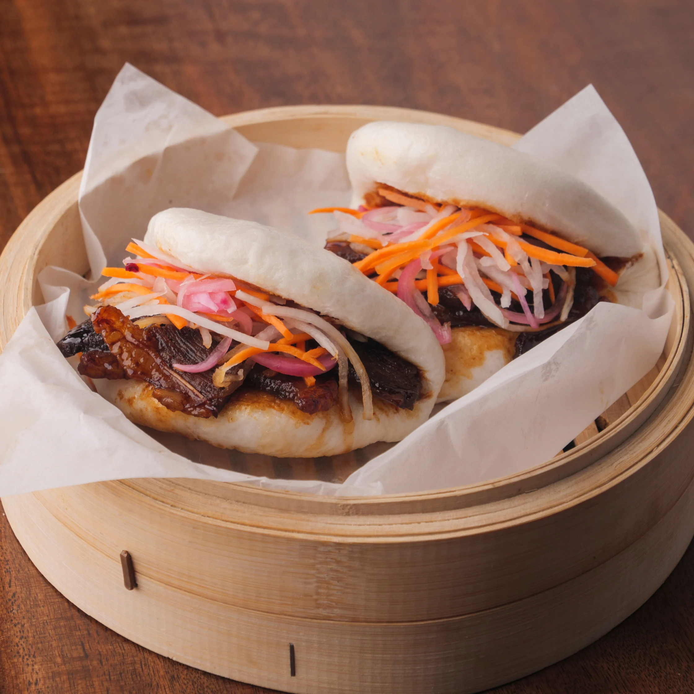 Two steamed bao buns filled with braised beef, topped with Filipino-style pickled vegetables (atchara) made from carrots, onions, and radish, served in a bamboo steamer basket.