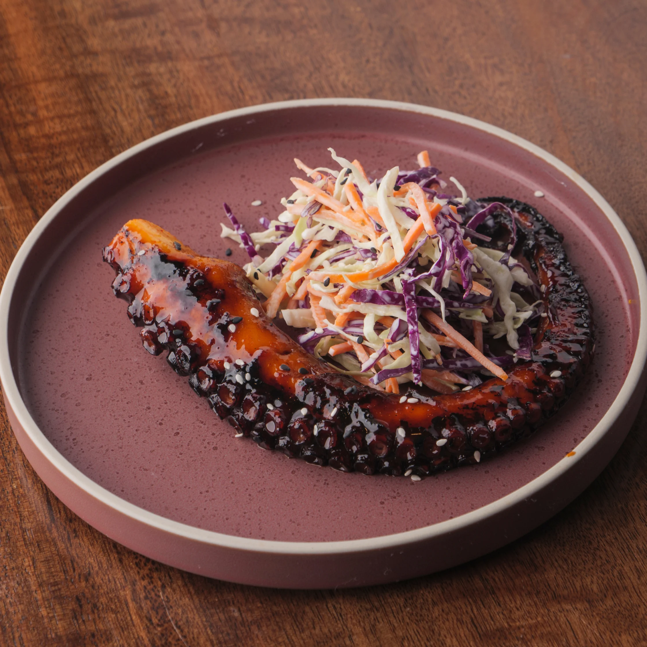 A grilled octopus tentacle glazed with a dark sauce and sprinkled with sesame seeds, plated with a colorful cabbage slaw on a mauve ceramic dish.