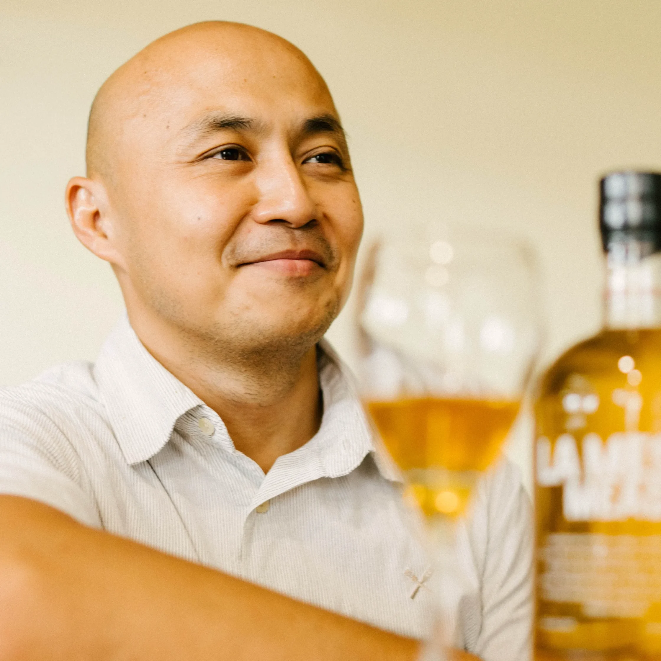 ortrait of the LA MESA MEAD owner smiling while holding a glass of golden mead, with a branded bottle softly blurred in the foreground.