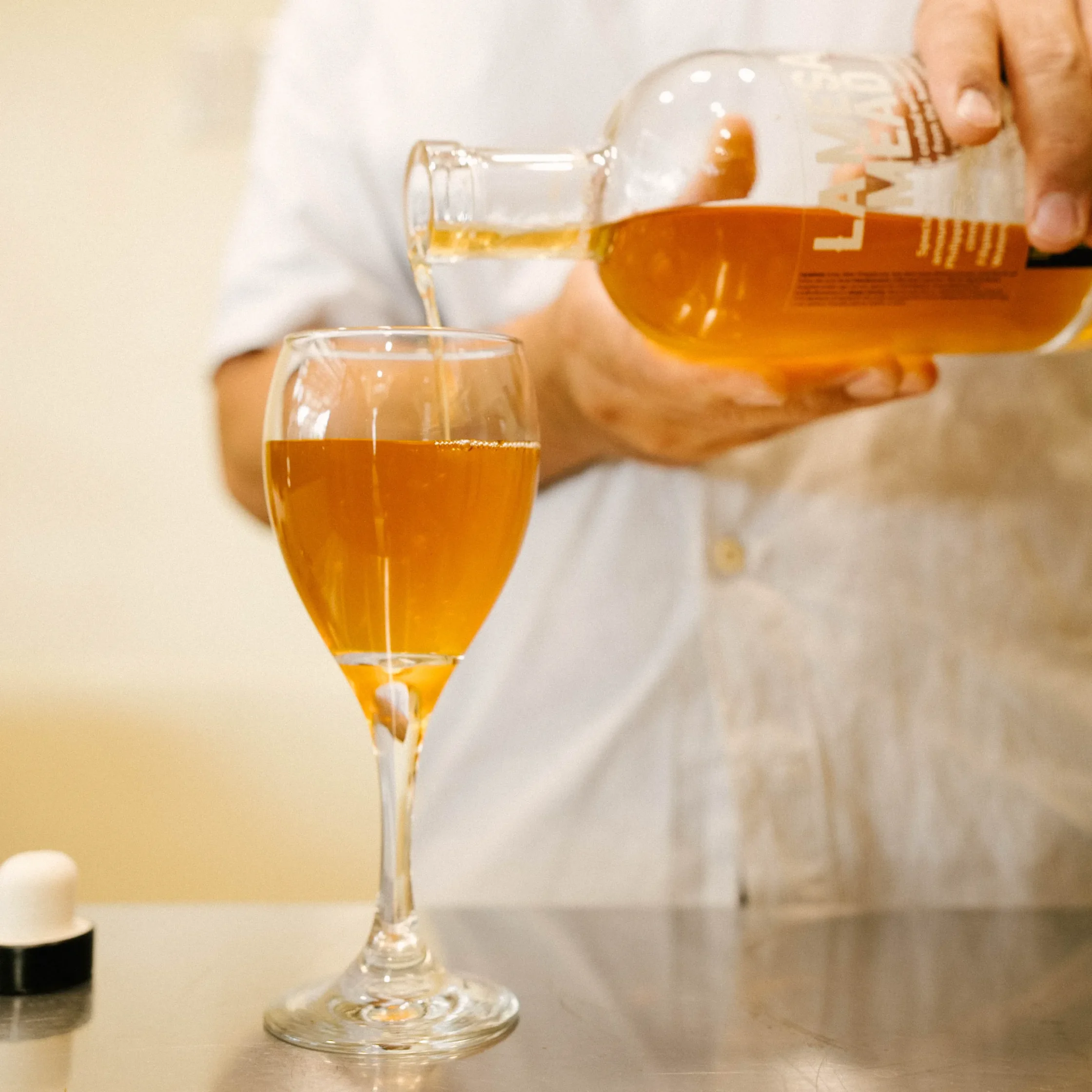 Golden LA MESA MEAD being poured from a branded bottle into a wine glass, highlighting the beverage’s clear amber color.