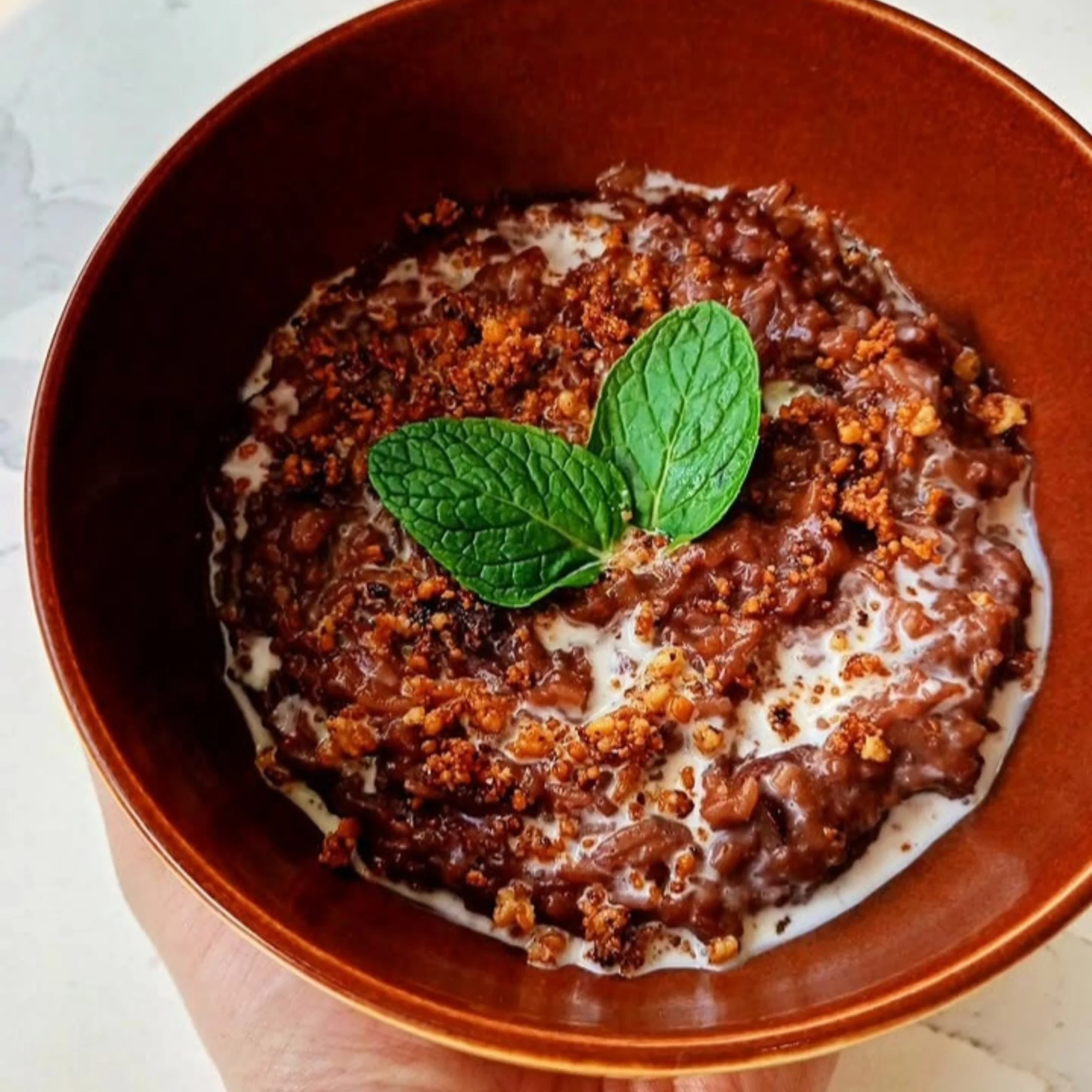 Bowl of rich chocolate champorado topped with crunchy toasted crumbs and fresh mint leaves, finished with a drizzle of cream.