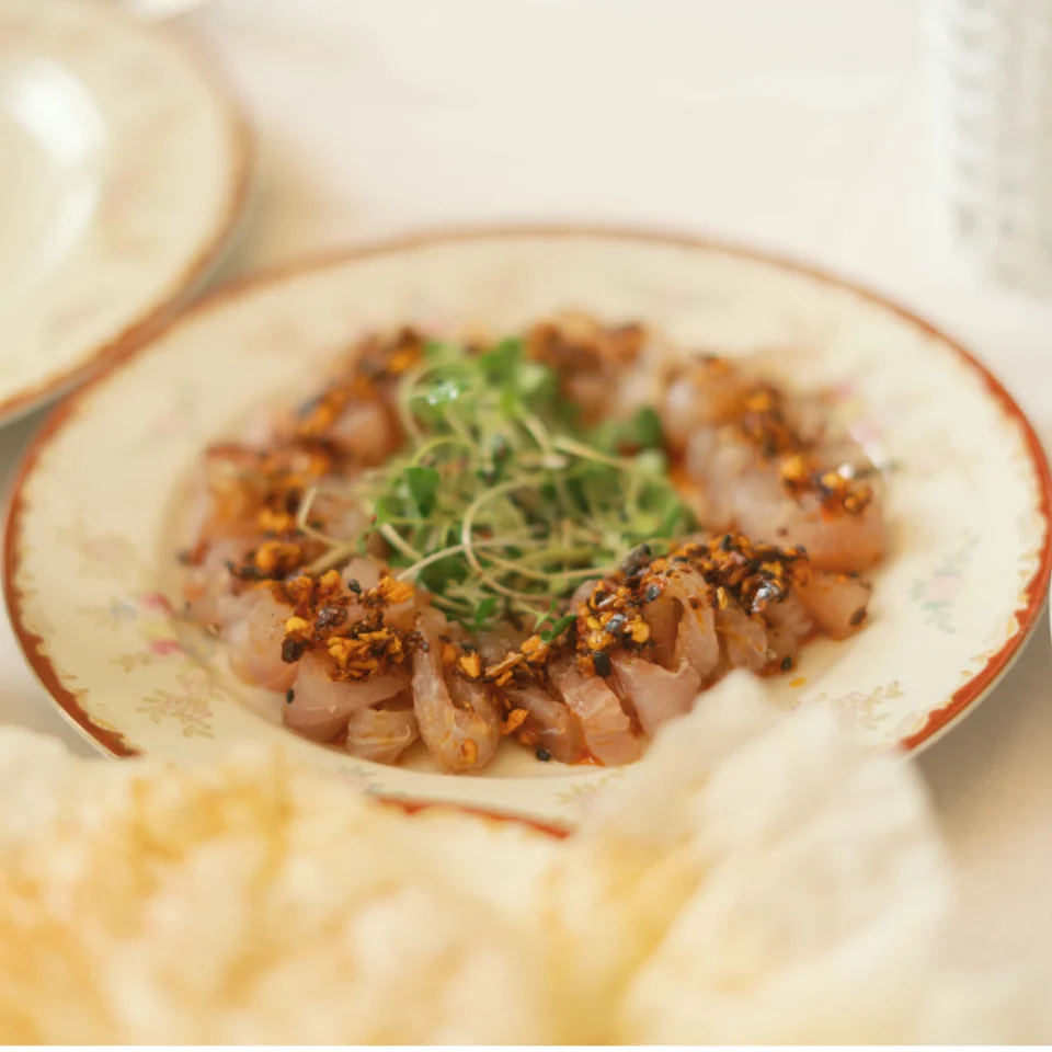 Thin slices of five-spice cured sea bream arranged on a decorative plate, topped with chili-garlic seasoning and fresh microgreens.