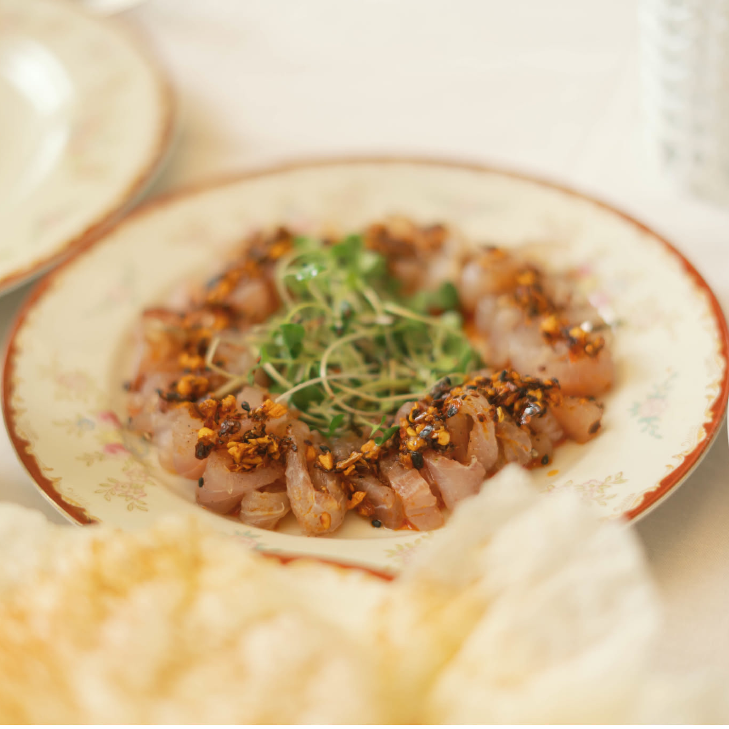 Thin slices of five-spice cured sea bream arranged on a decorative plate, topped with chili-garlic seasoning and fresh microgreens.