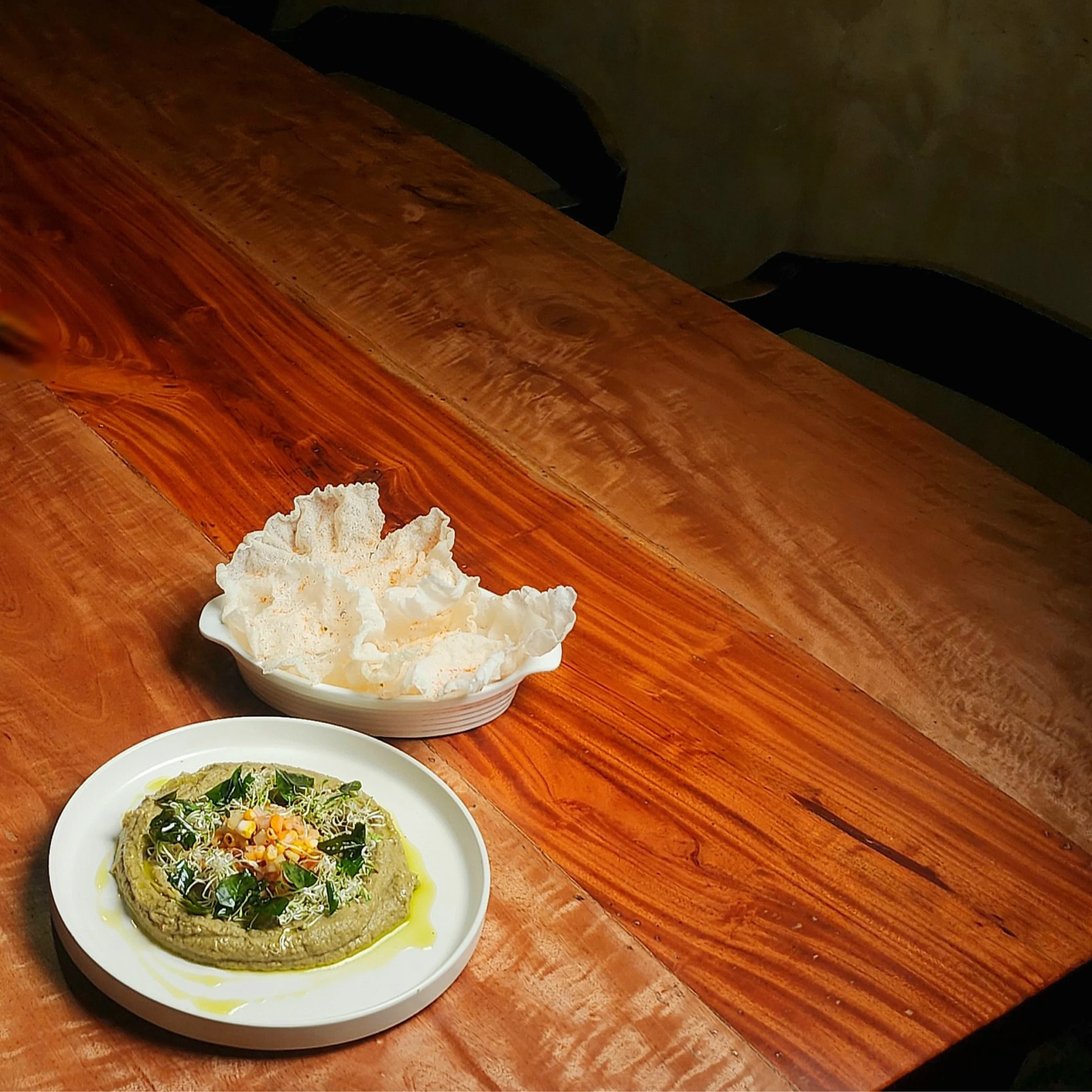 Plate of hummus verde garnished with herbs and olive oil, served alongside a bowl of crispy prawn crackers on a wooden table.