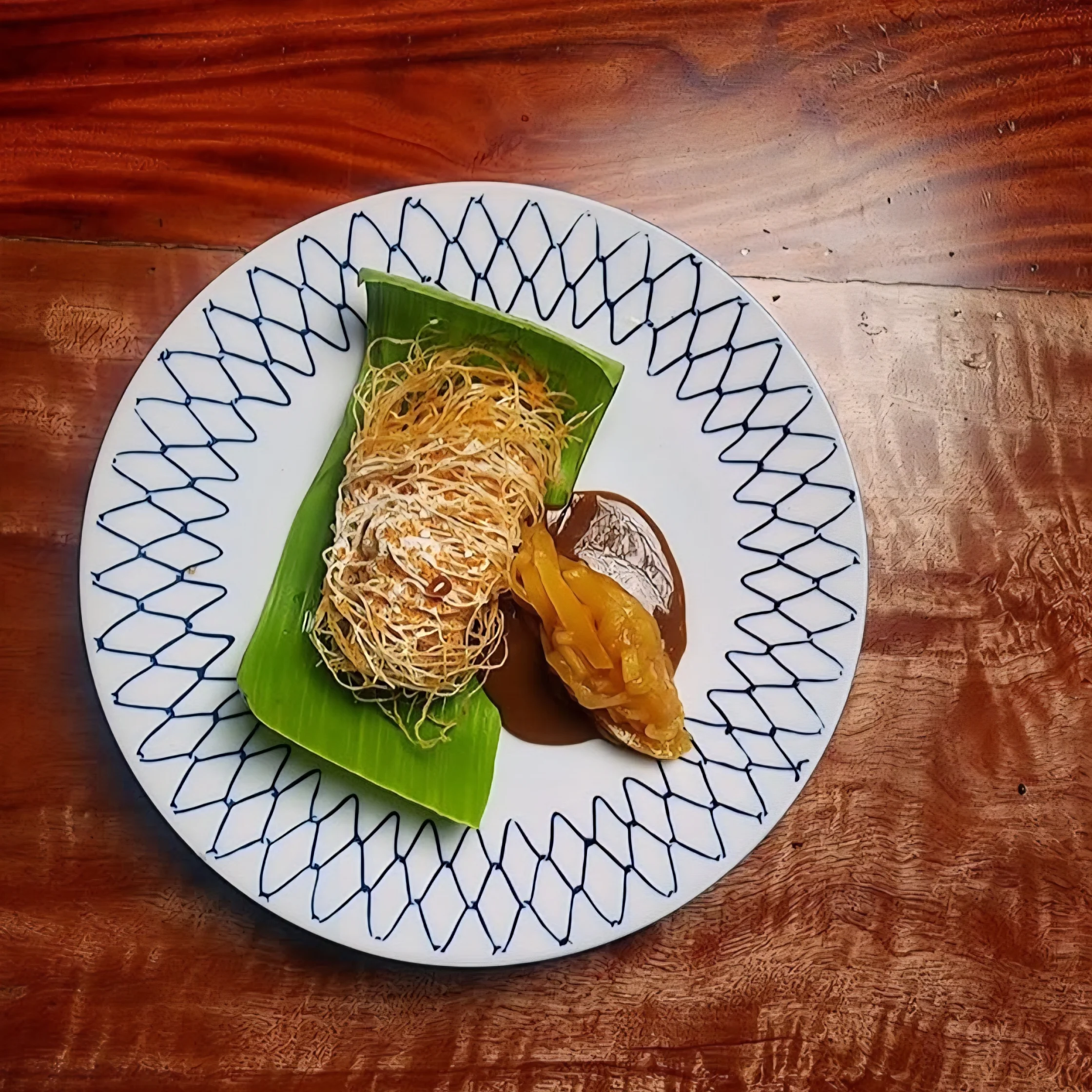 A plated dessert featuring a crisp, nest-like fried pastry roll resting on a banana leaf, dusted with powdered sugar and served with caramel-colored sauce and a piece of fruit on a patterned white plate atop a wooden table.