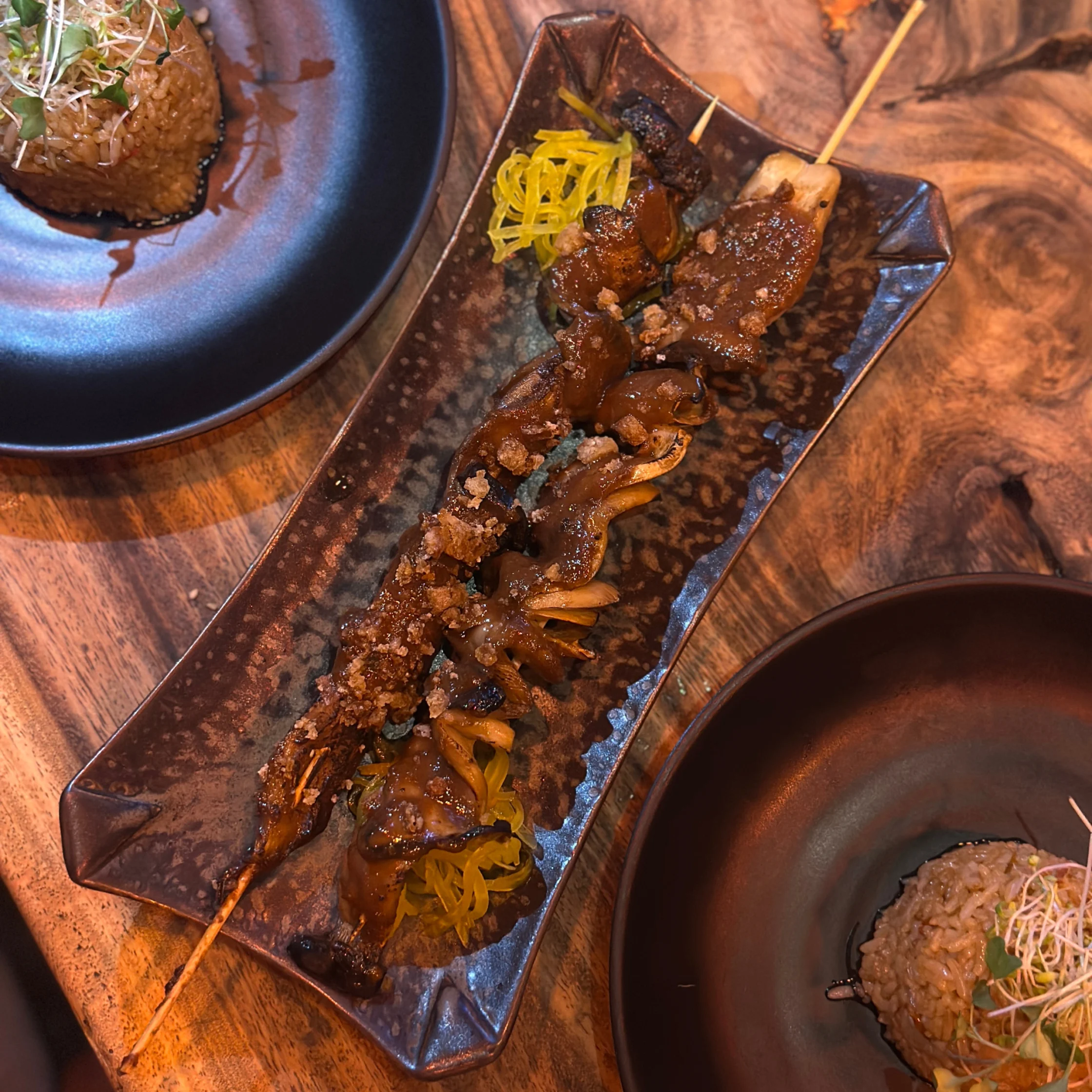 Skewers of glazed grilled mushrooms arranged on a long dark ceramic platter, topped with crispy bits and served with pickled vegetables, surrounded by other plated dishes on a wooden dining table.