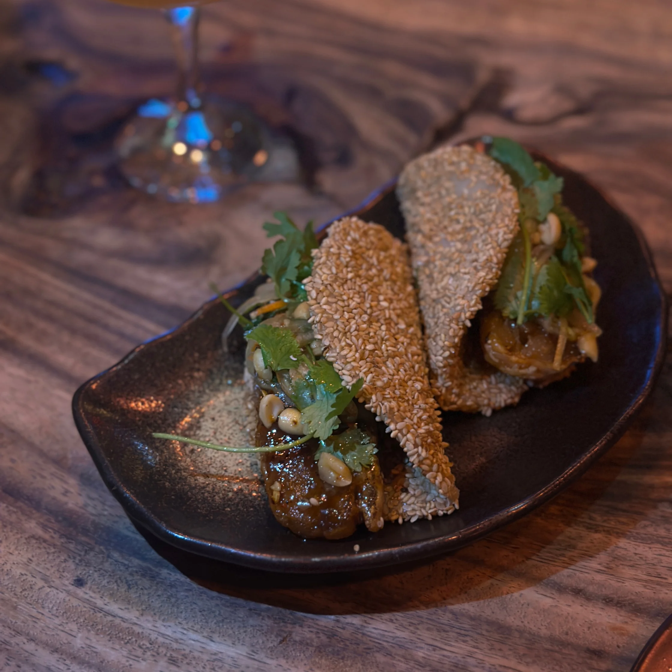 Two sesame-crusted bao-style buns filled with savory meat, herbs, and vegetables, presented on a dark plate with a blurred wine glass in the background.