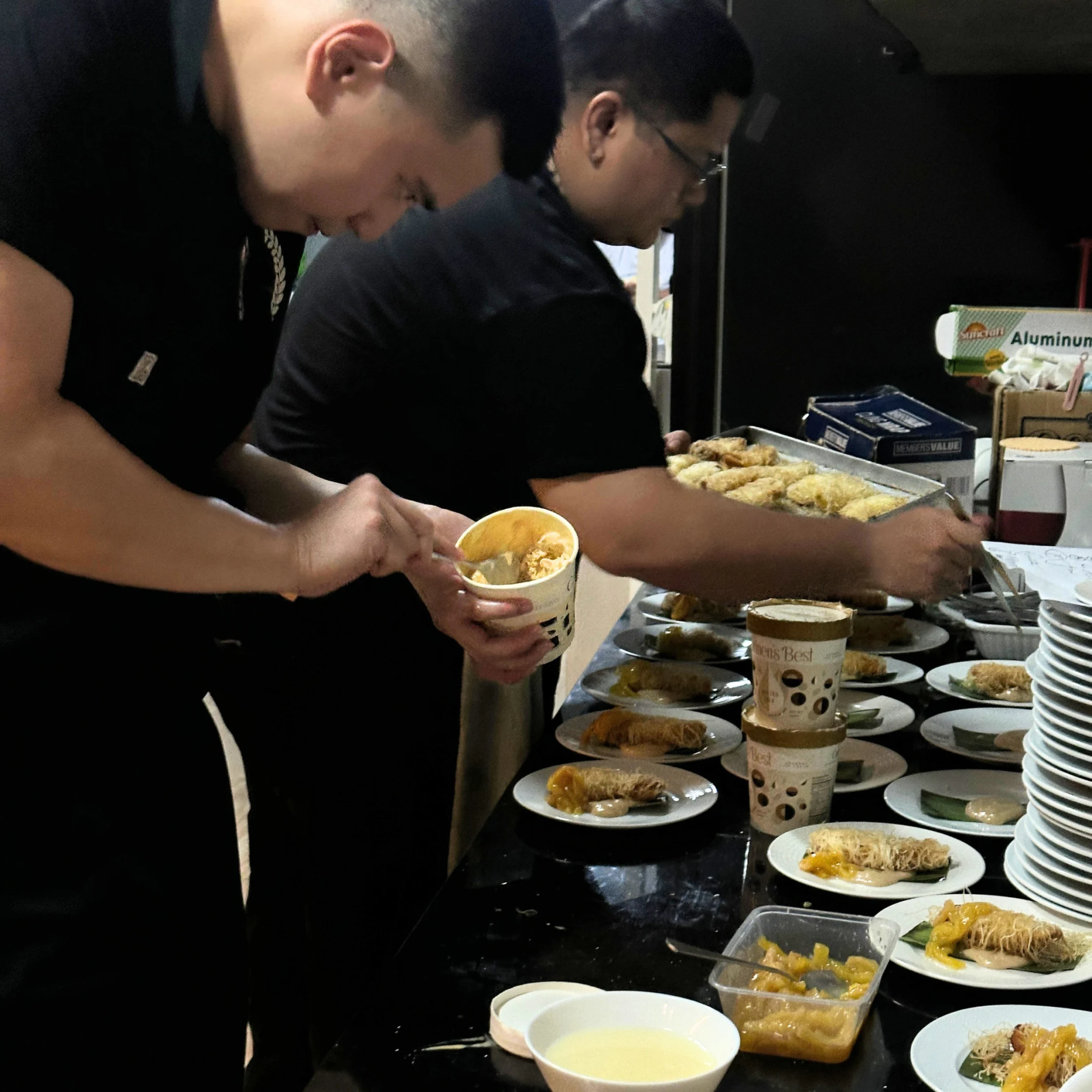 Two cooks plating multiple dishes in a kitchen or dining prep area, arranging desserts and garnishes on rows of plates set across a counter.