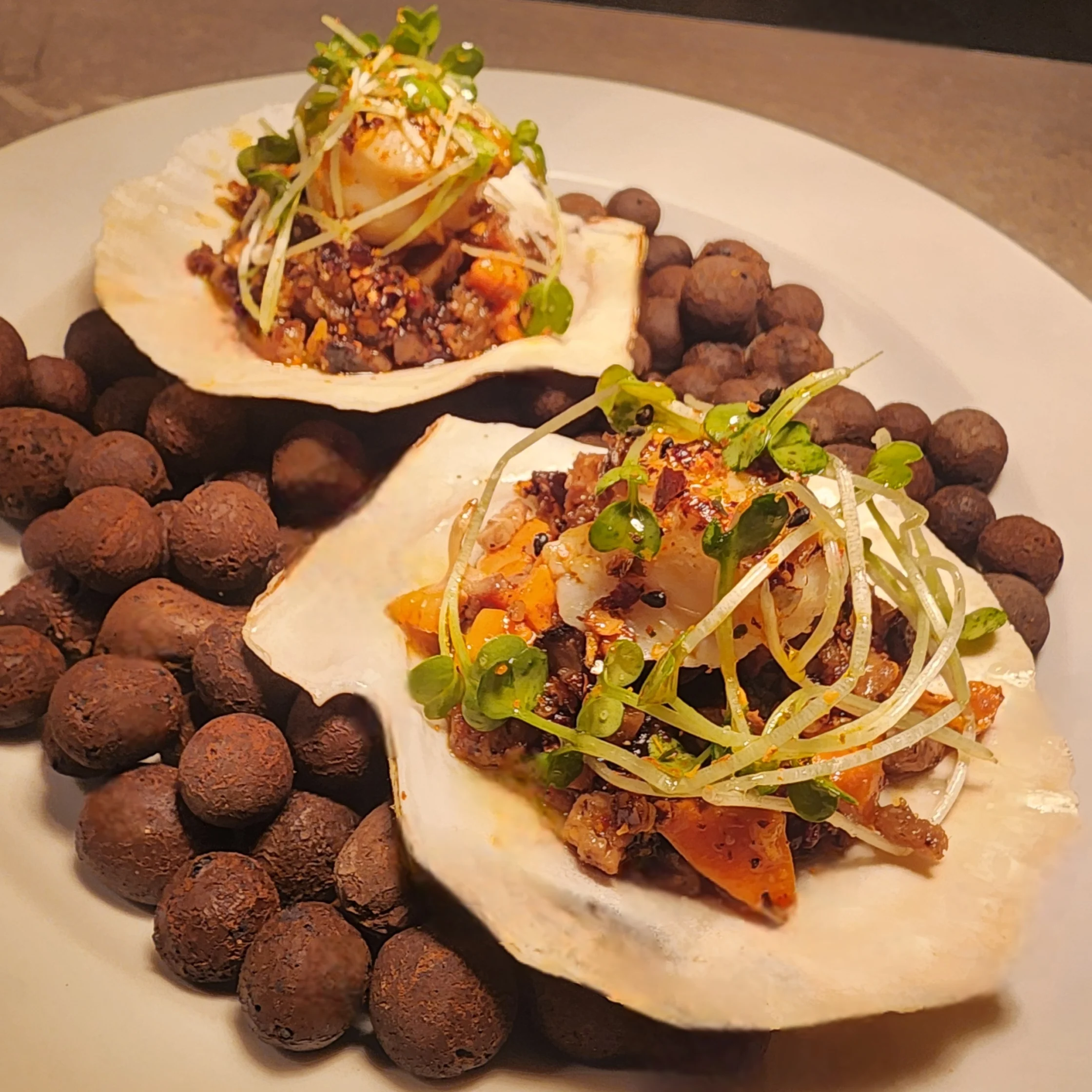 Two large shell halves filled with a finely chopped seafood mixture, garnished with microgreens and spices, displayed on a plate surrounded by small dark decorative stones.