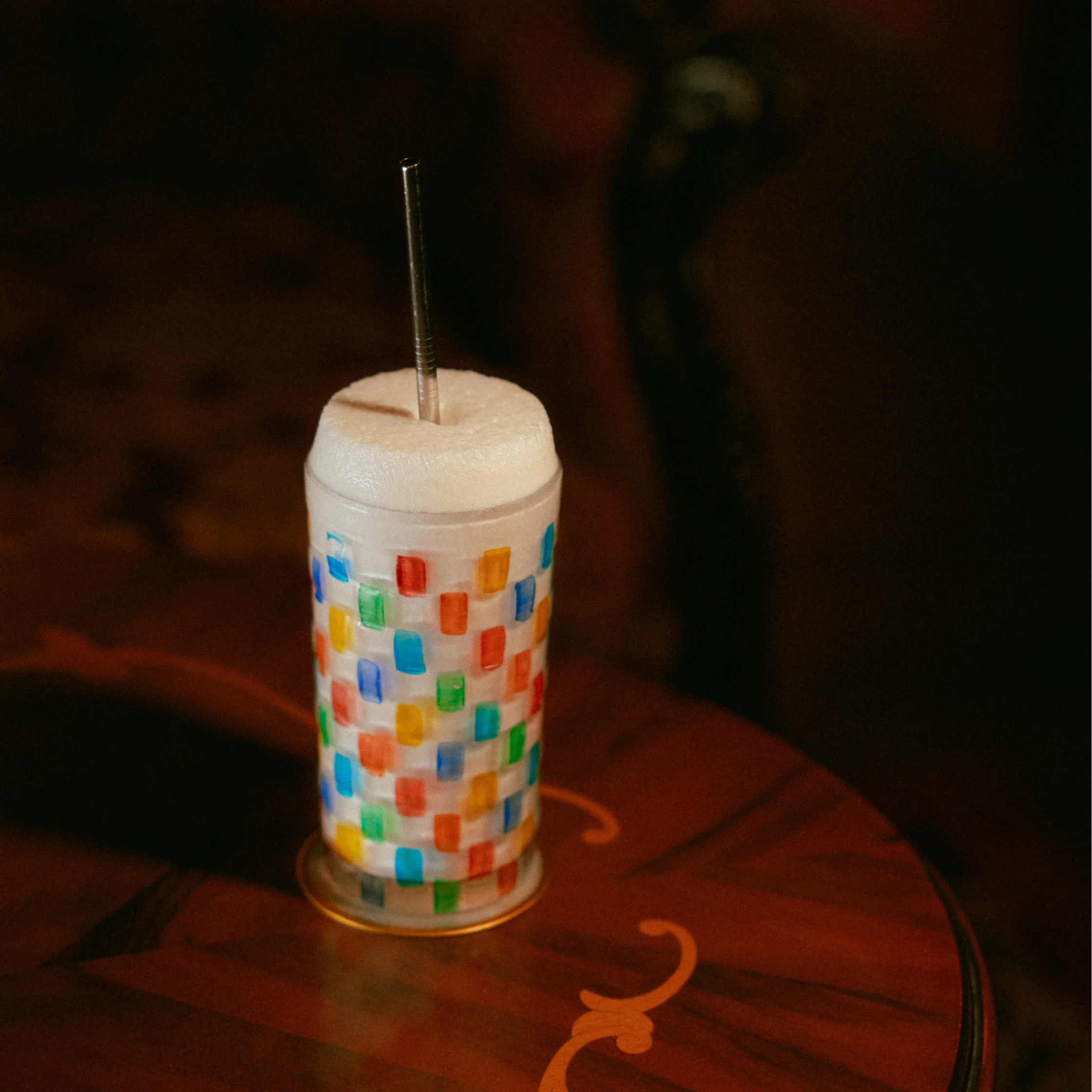 Tall frothy cocktail served in a colorful patterned glass with a metal straw, photographed on a dimly lit wooden table.
