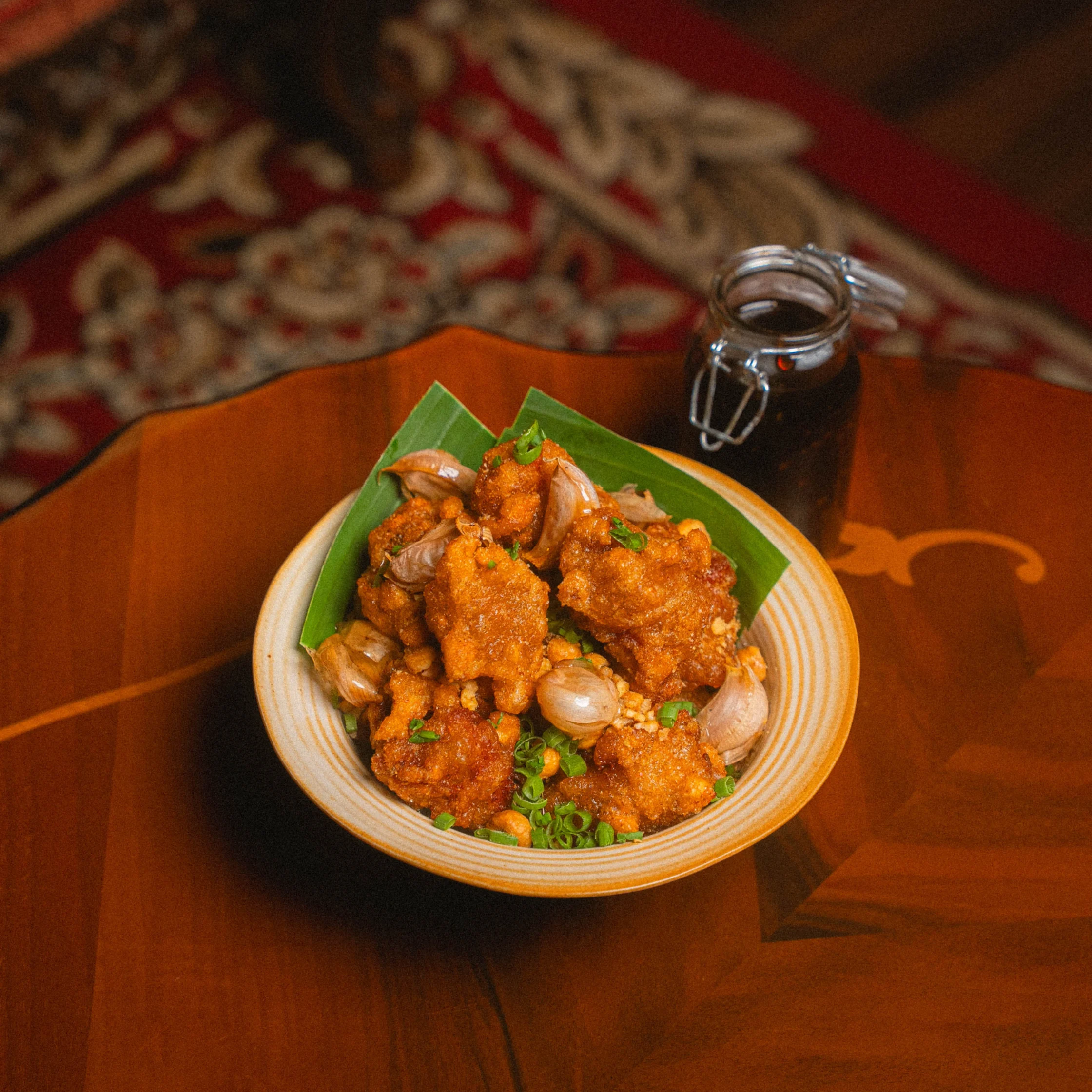 Bowl of crispy fried bites topped with roasted garlic cloves, chopped scallions, and crunchy garnish, served on banana leaves with a small bottle of dark dipping sauce on a polished wooden table.