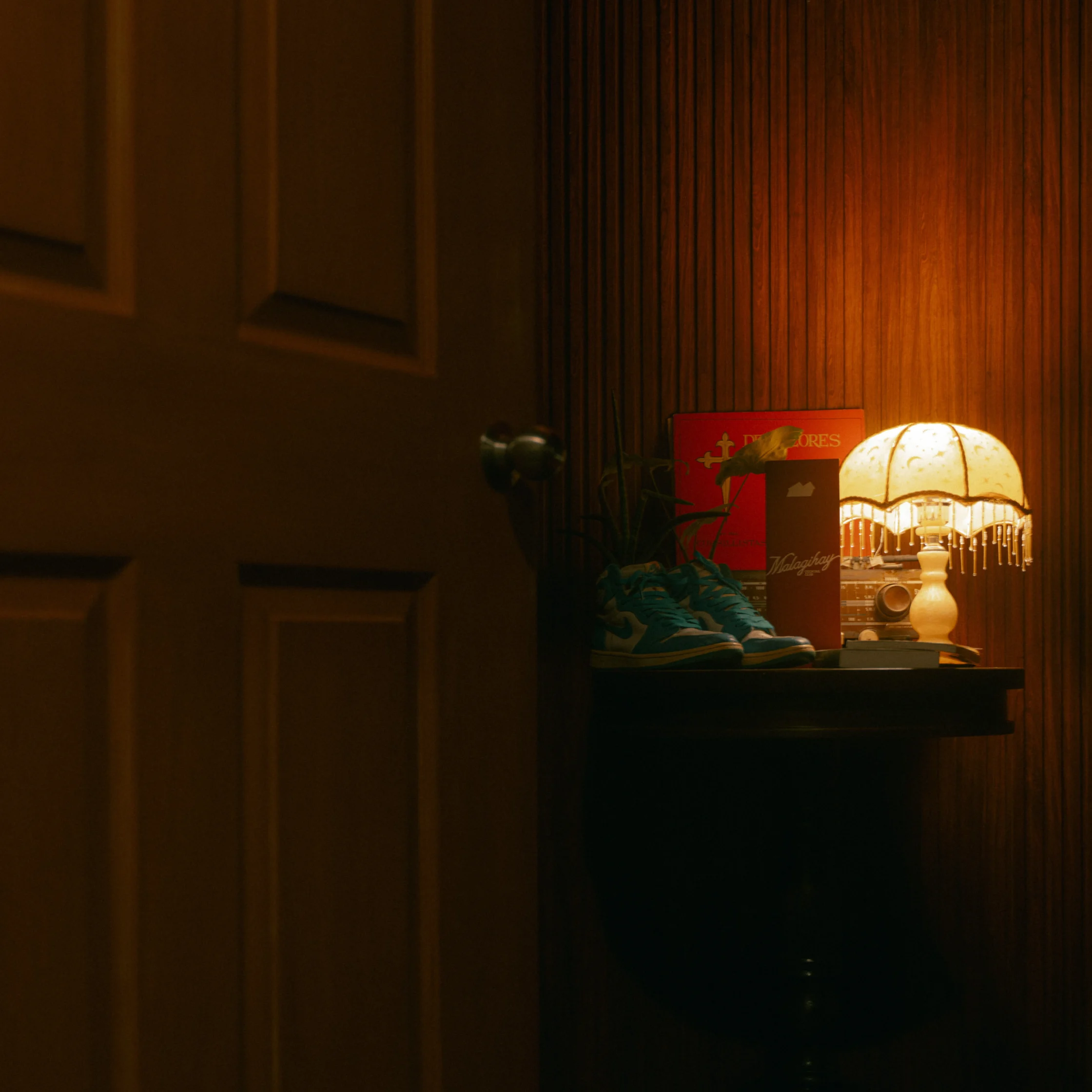 Warmly lit corner vignette featuring a vintage-style table lamp, framed items, sneakers, and decorative objects against dark wood paneling.