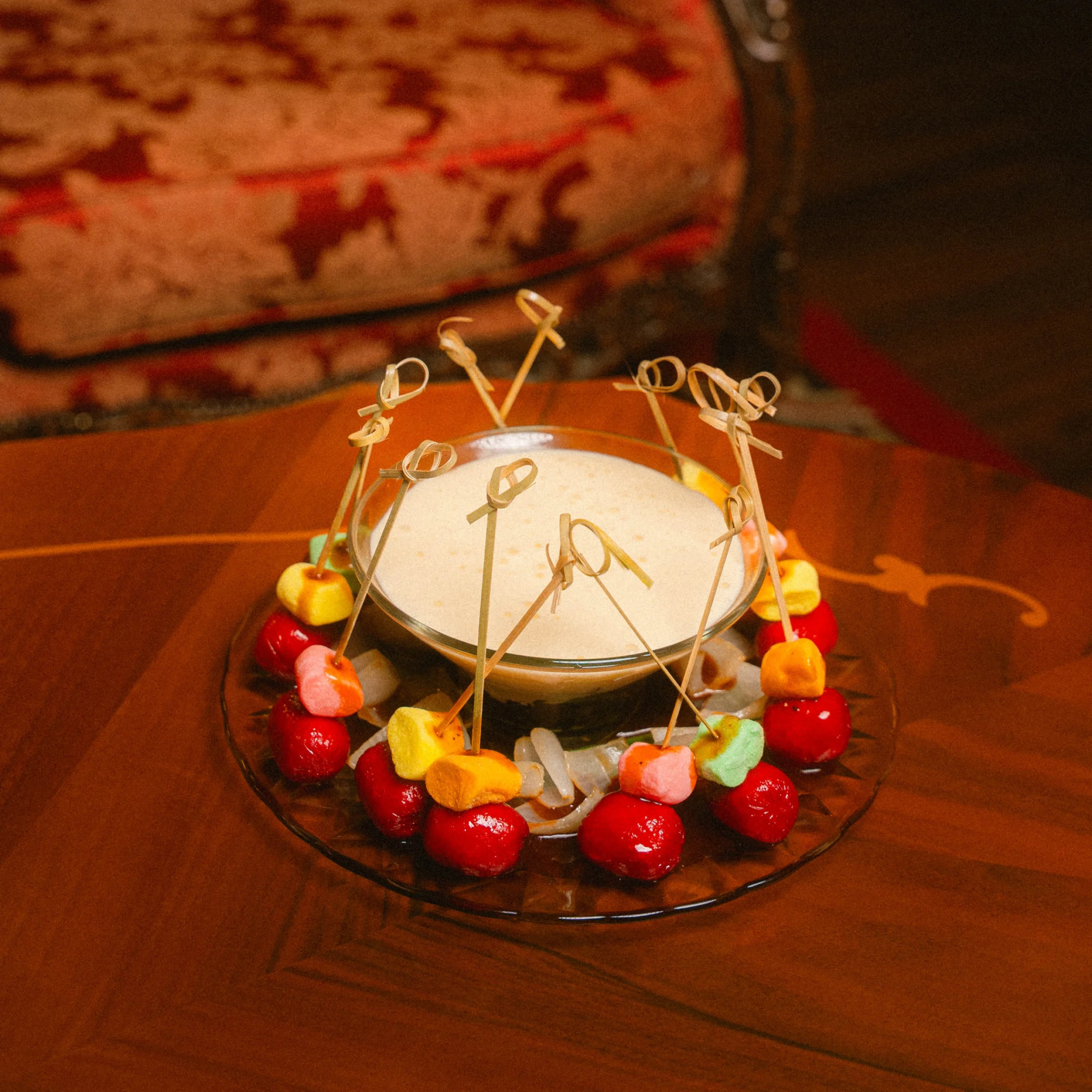 Glass bowl of creamy dip surrounded by skewers holding colorful bite-sized pieces and bright red cocktail garnishes arranged on a glass plate.