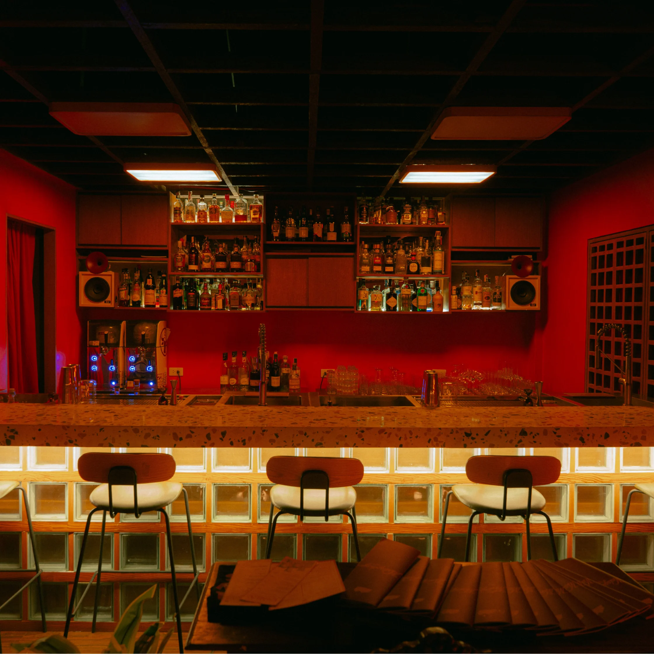 Warm red-toned interior of the cocktail club featuring a backlit bar, bottle displays, and seating in a cozy lounge atmosphere.