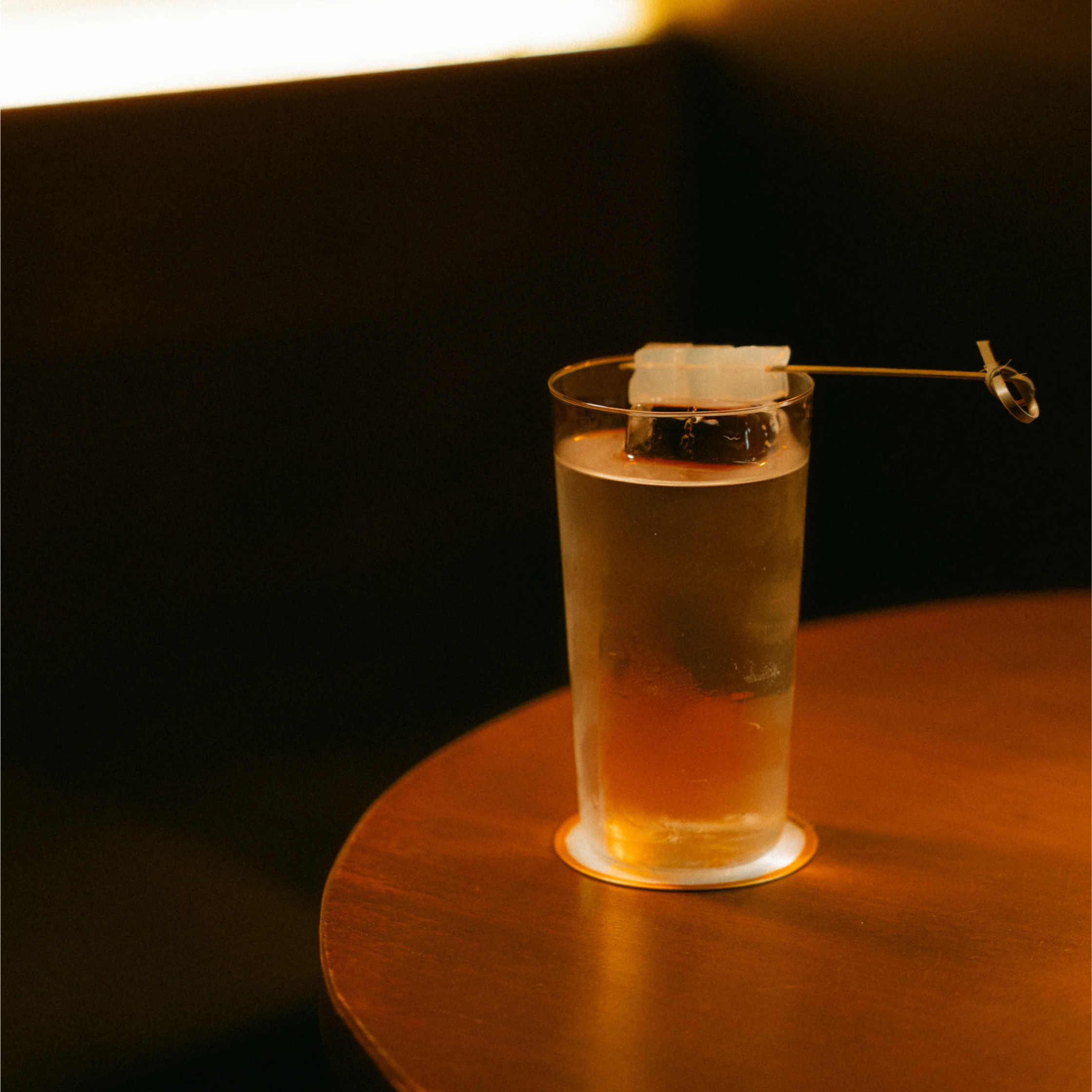 Clear layered cocktail served over ice in a tall glass with a decorative skewer, placed on a round wooden table.