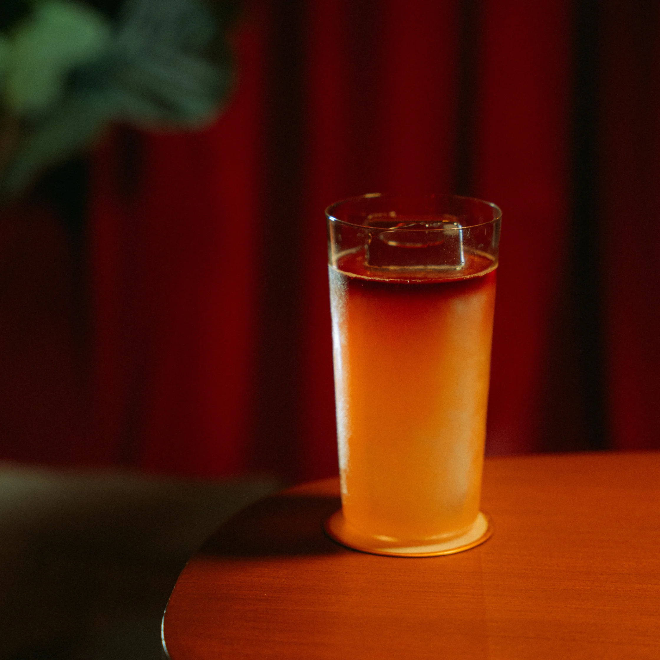 Tall chilled cocktail with a subtle gradient and large clear ice cube, set against red curtains and soft ambient lighting.