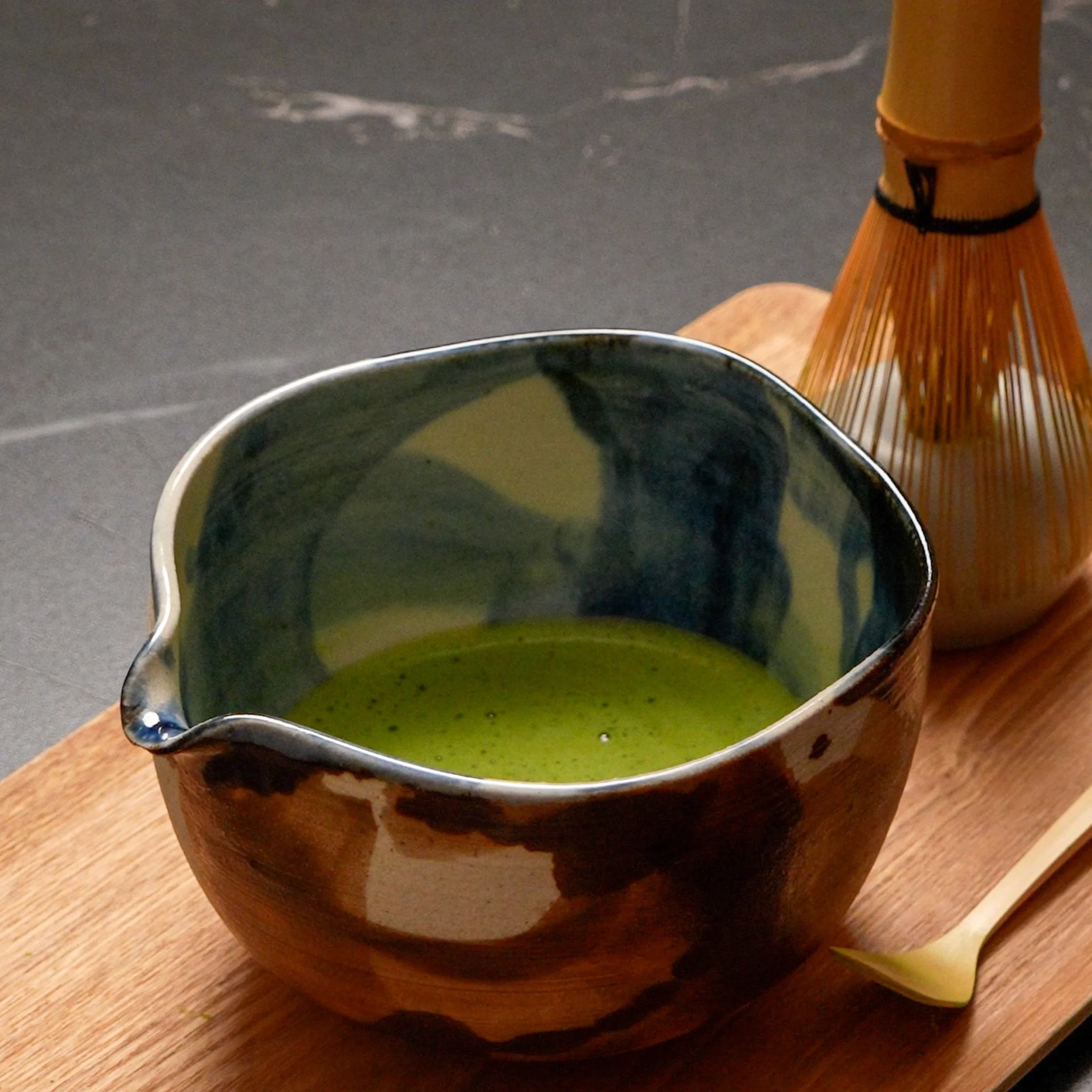 A bowl of freshly whisked matcha sits on a wooden tray beside a traditional bamboo whisk and scoop, ready for serving.