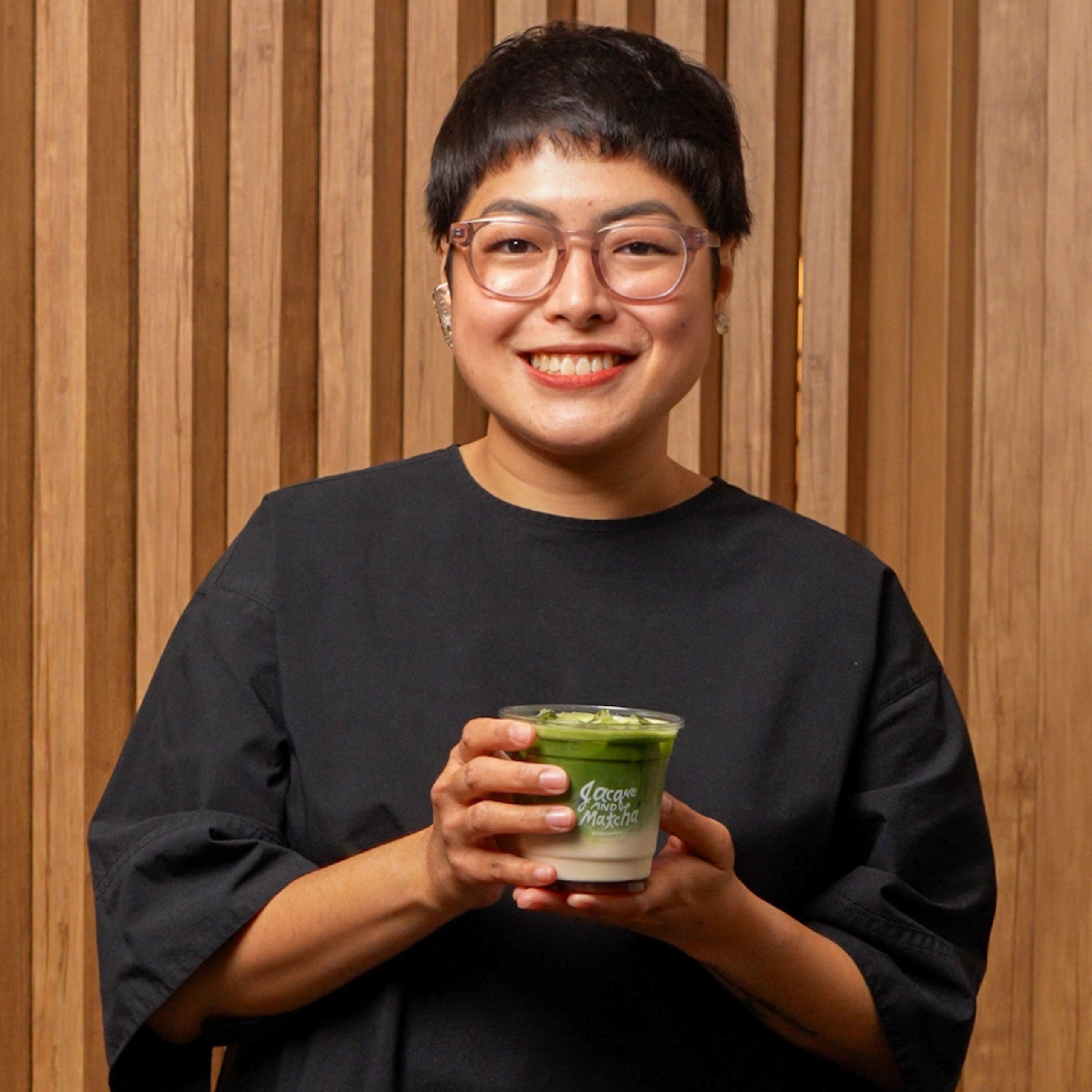 Jacque de Borja-Medestomas smiles while holding an iced matcha latte in front of a warm wooden backdrop, presenting her signature matcha beverage.