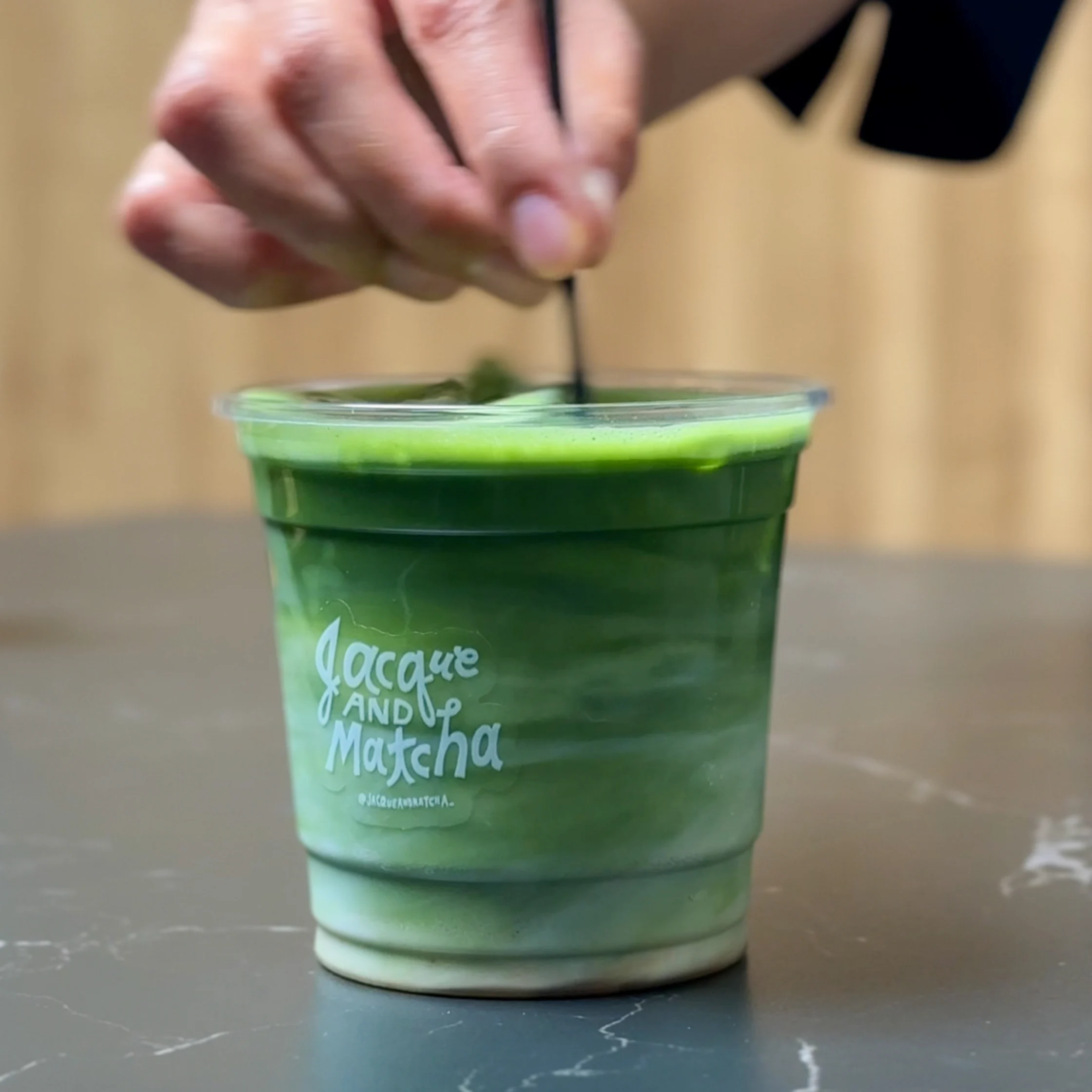 A close-up of a hand stirring an iced matcha latte in a clear “Jacque and Matcha” cup, blending the layers of milk and matcha.