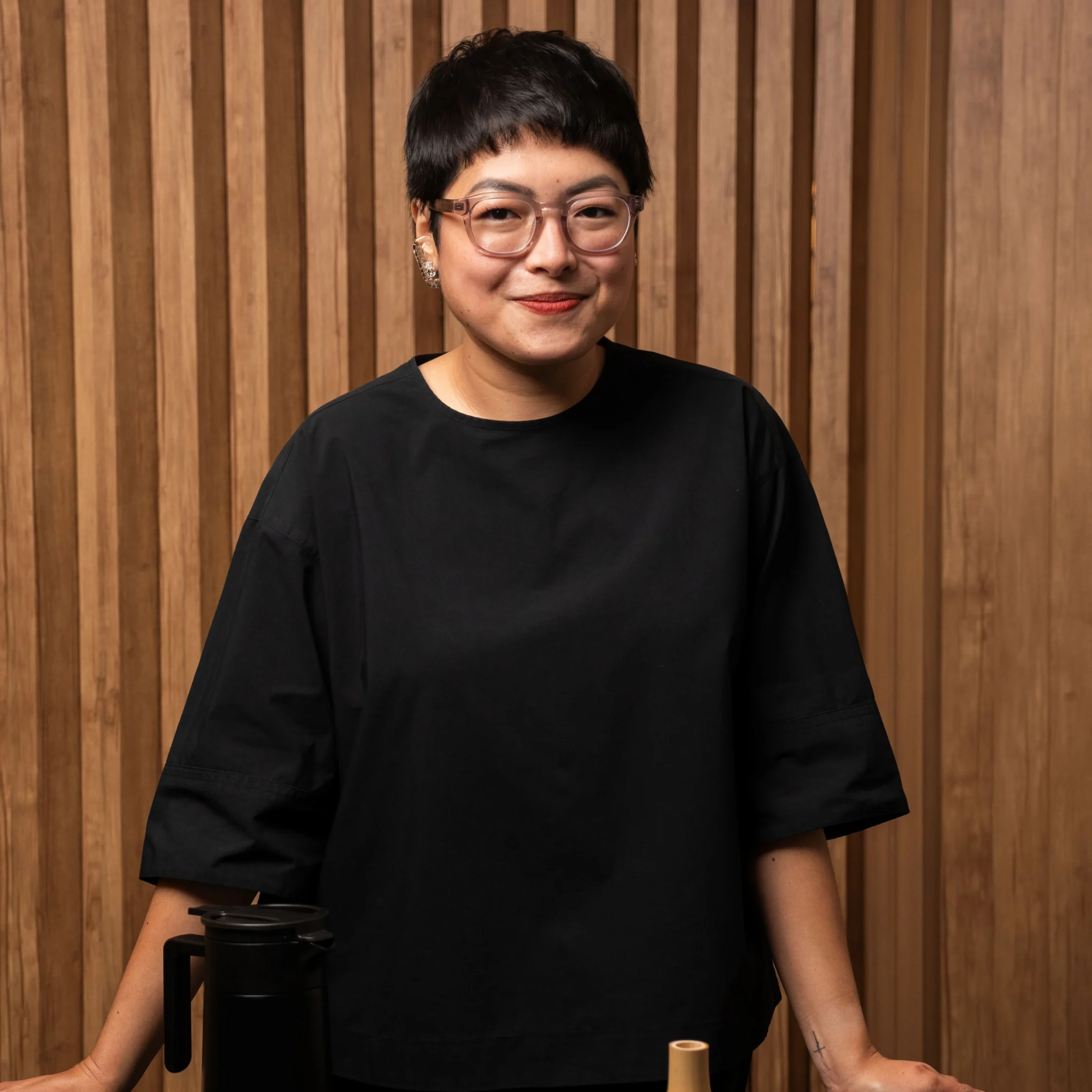 Jacque de Borja-Medestomas stands in front of a wooden-paneled wall, smiling at the camera while posing beside matcha-making equipment, including a kettle and traditional bamboo whisk, during a matcha workshop.