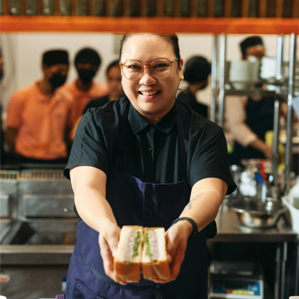 Miko Calo presenting a sandwich toward the camera from behind a restaurant counter.