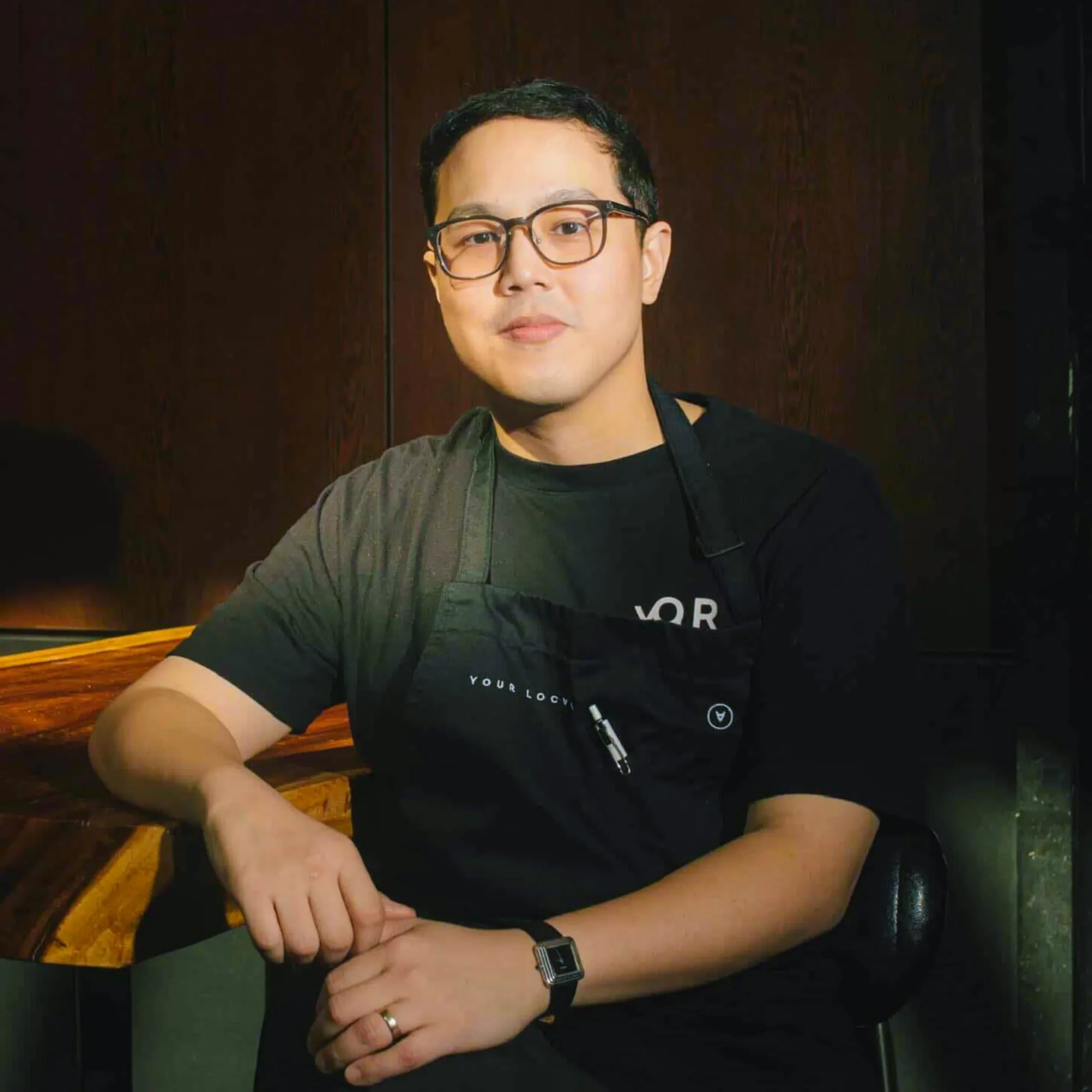 Chef Pat Go of Your Local wearing glasses, a black shirt, and an apron, seated beside a wooden counter in a dimly lit restaurant interior.