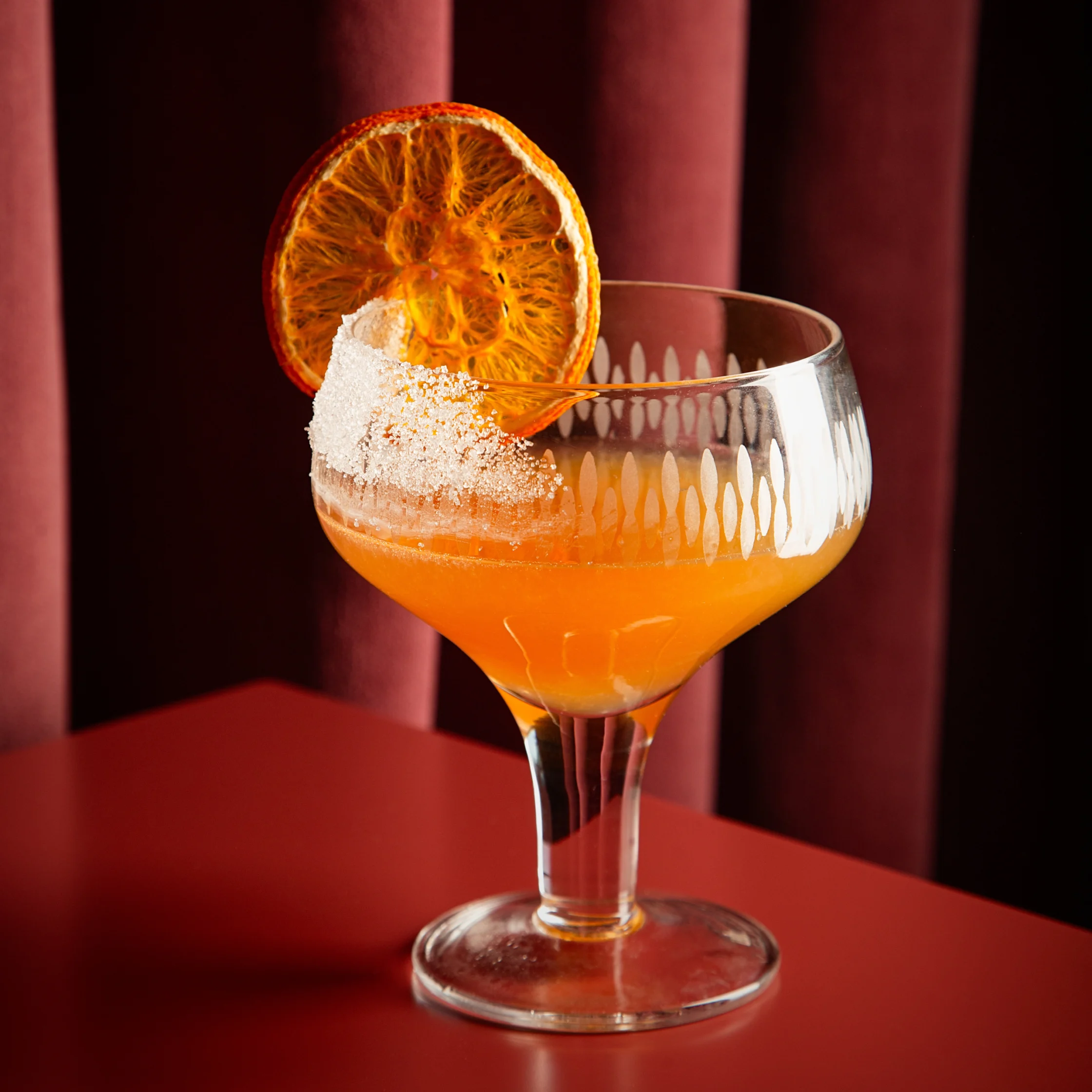 BRONTË Sidecar cocktail in a coupe glass, garnished with a dehydrated orange wheel and a mound of sugar, set against deep burgundy banquette seating.