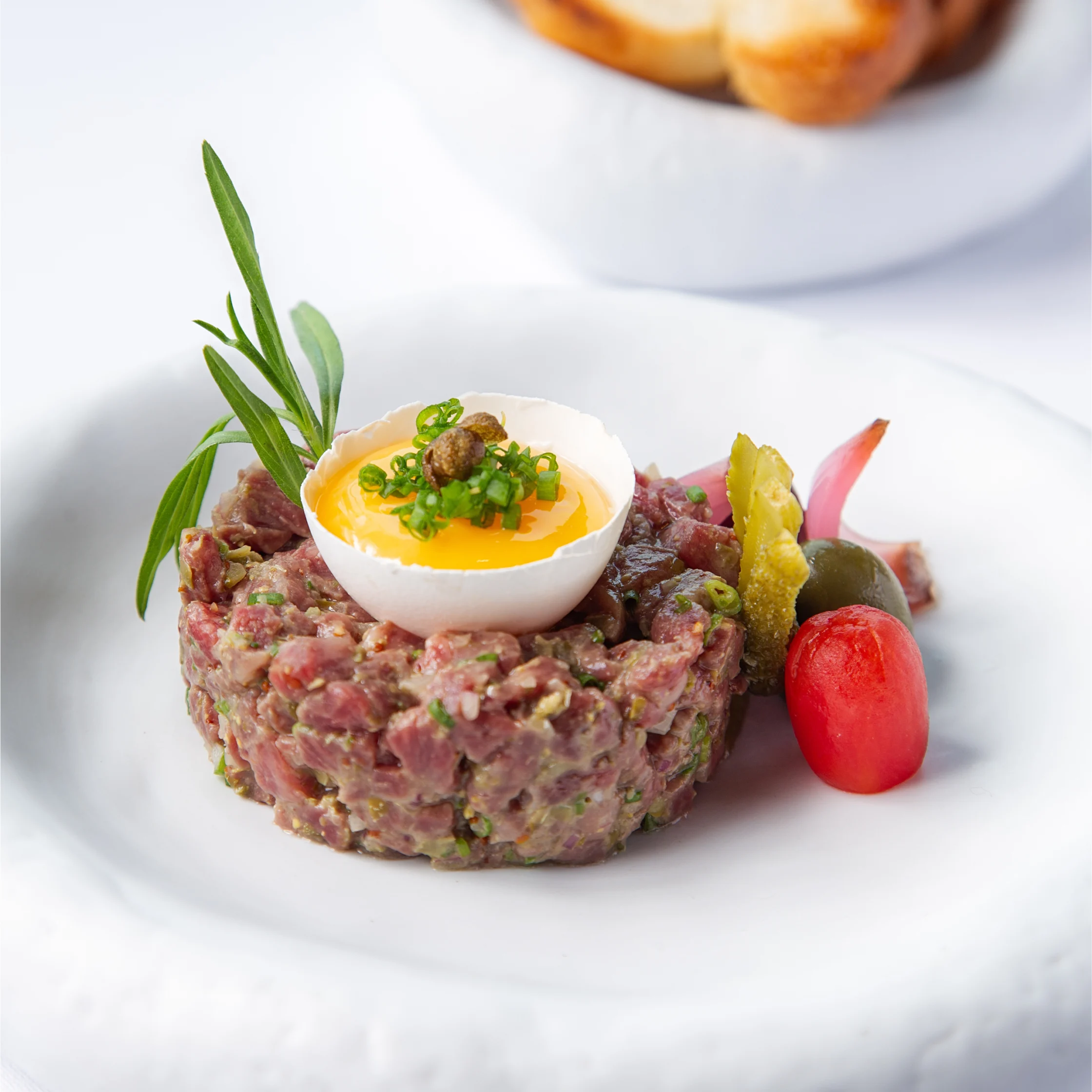 BRONTË Steak Tartare presented in a neat round mould, topped with an egg yolk in a half eggshell, chives, and capers, with pickles and a cherry tomato on the side.