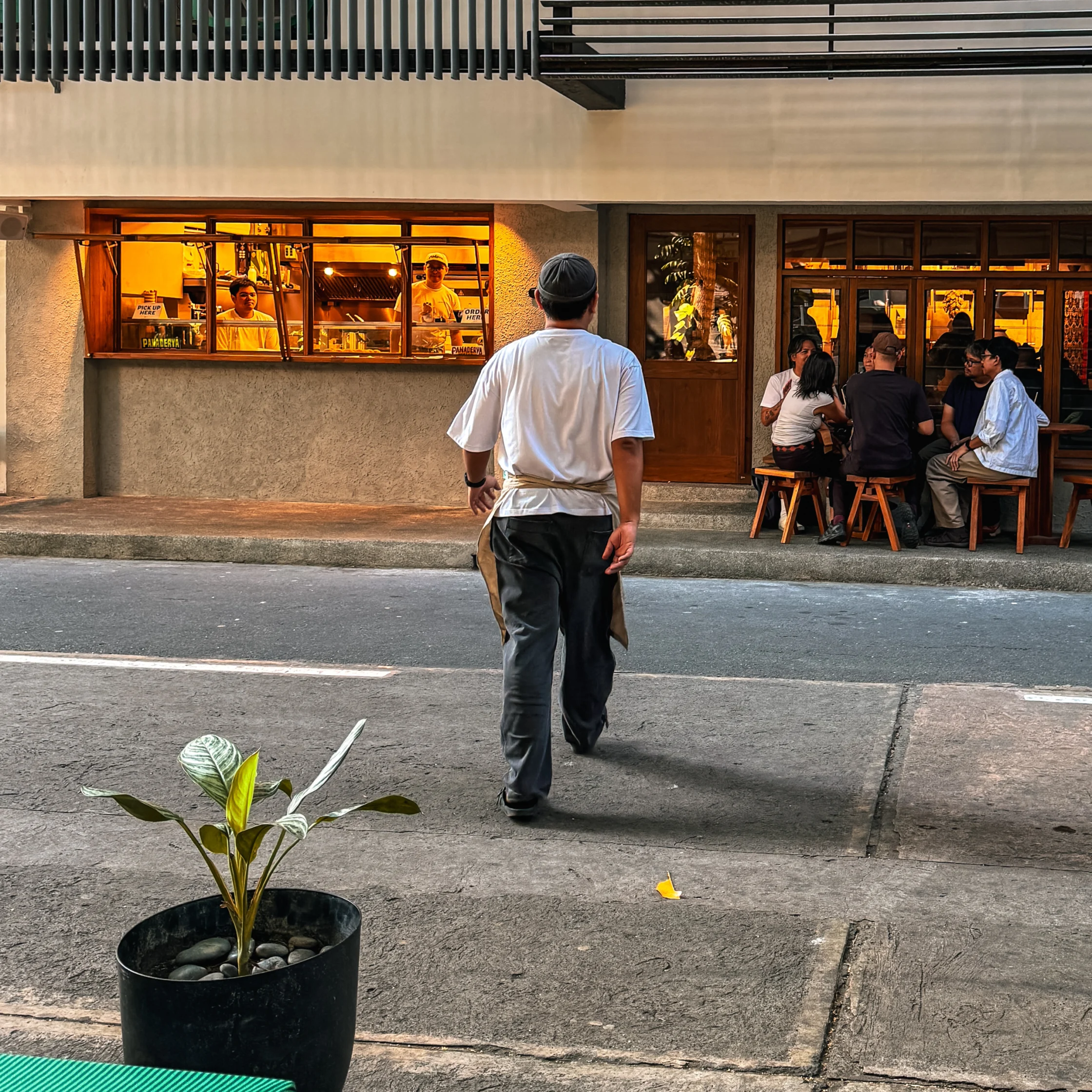 A bakery staff member walks across the street toward Toyo Panaderya’s new Karrivin, Makati location, with customers seated outside and warm light glowing from the bakery windows.