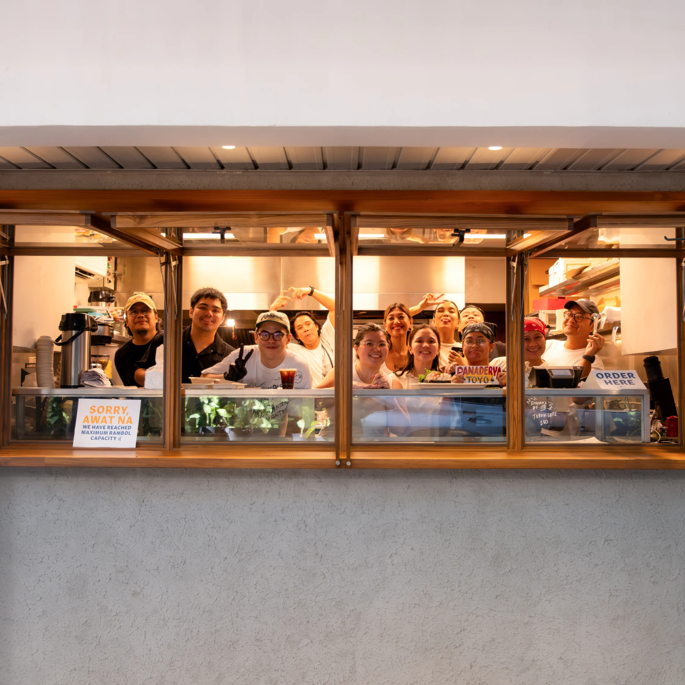 Group portrait of the Toyo Panaderya team smiling and posing together behind the bakery’s wooden-framed service window.