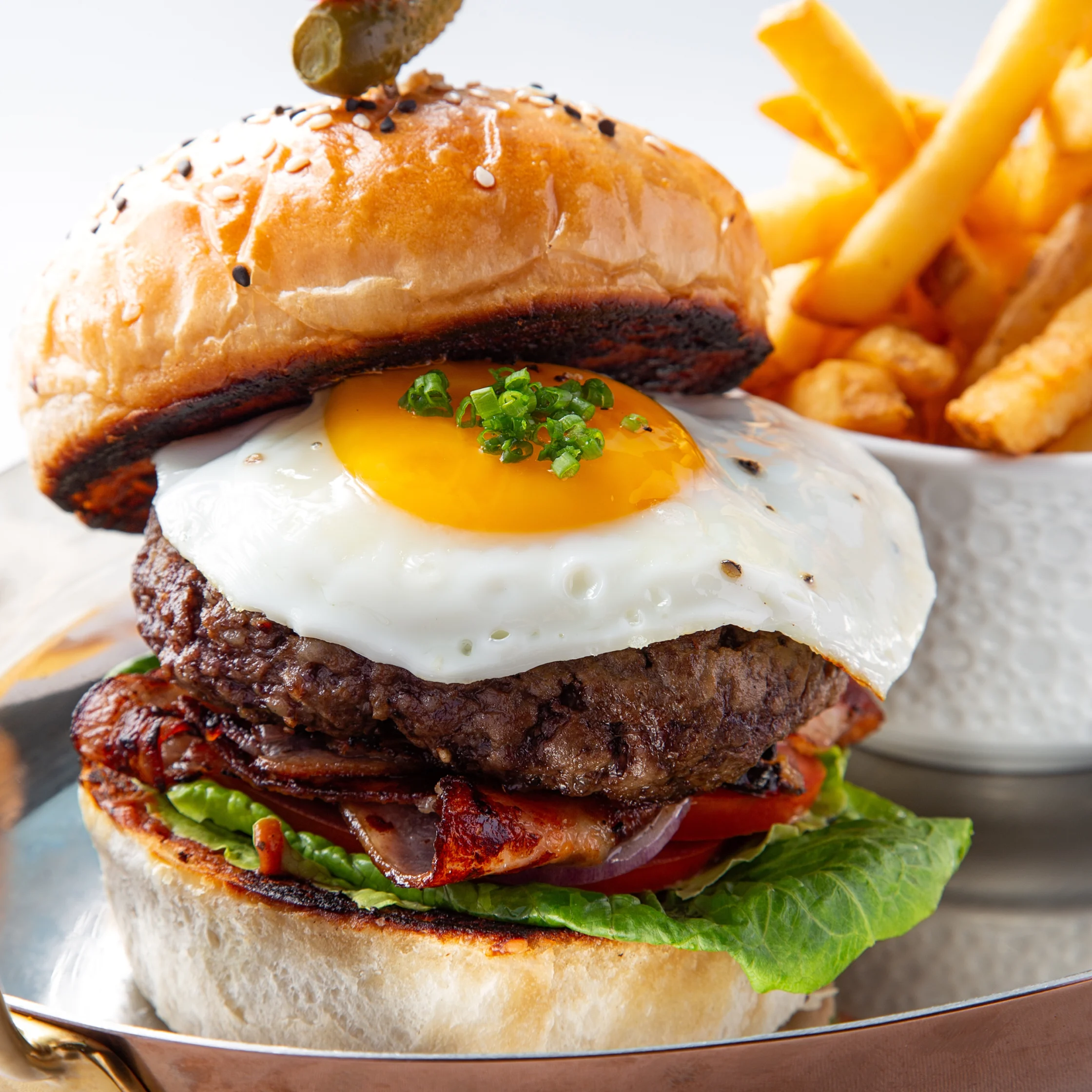 BRONTË Burger stacked with a beef patty, fried egg, bacon, lettuce, tomato, and onions in a toasted sesame bun, served with golden fries.