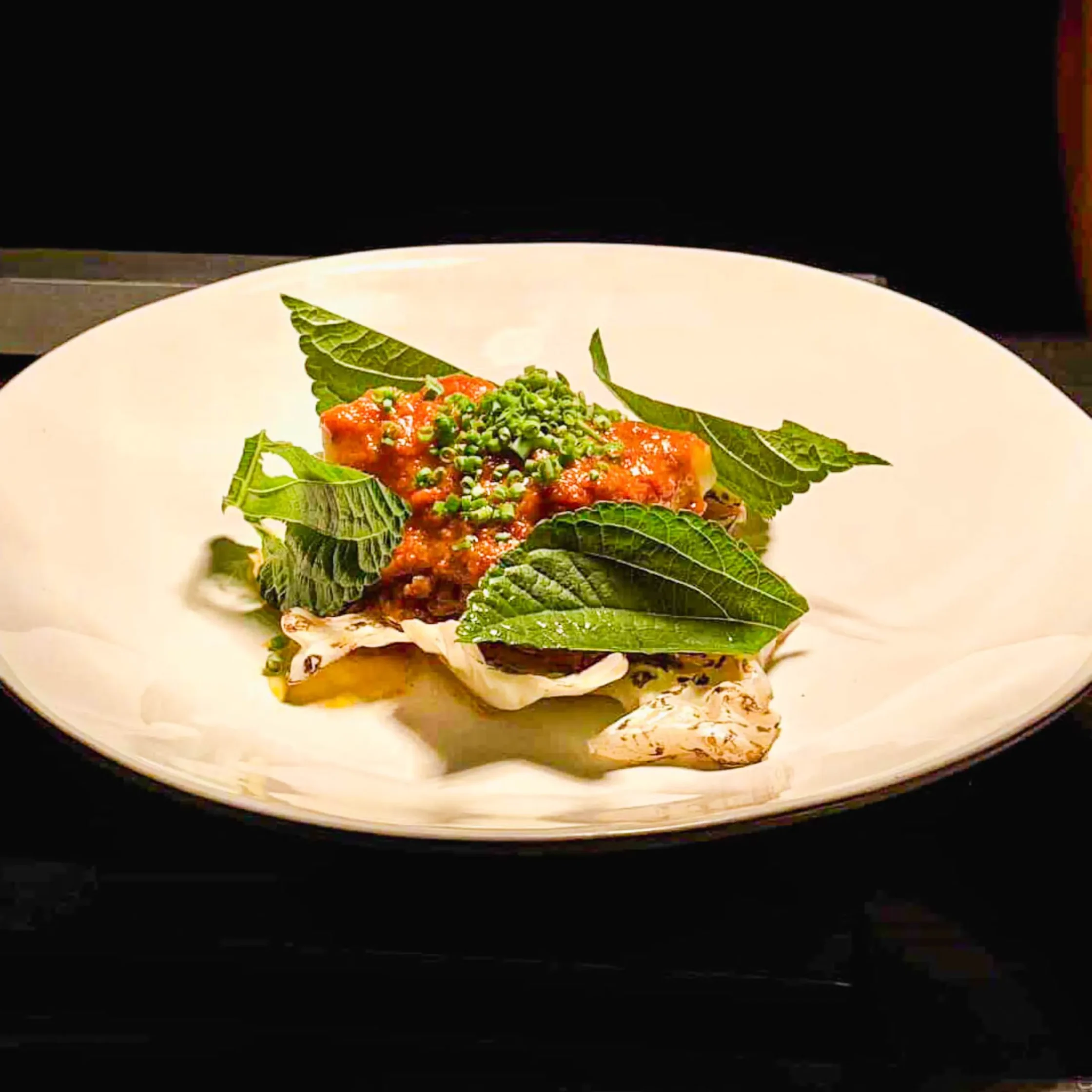 Plated halibut dish topped with red rendang-style sauce, fresh green leaves, and chopped herbs on a white plate.