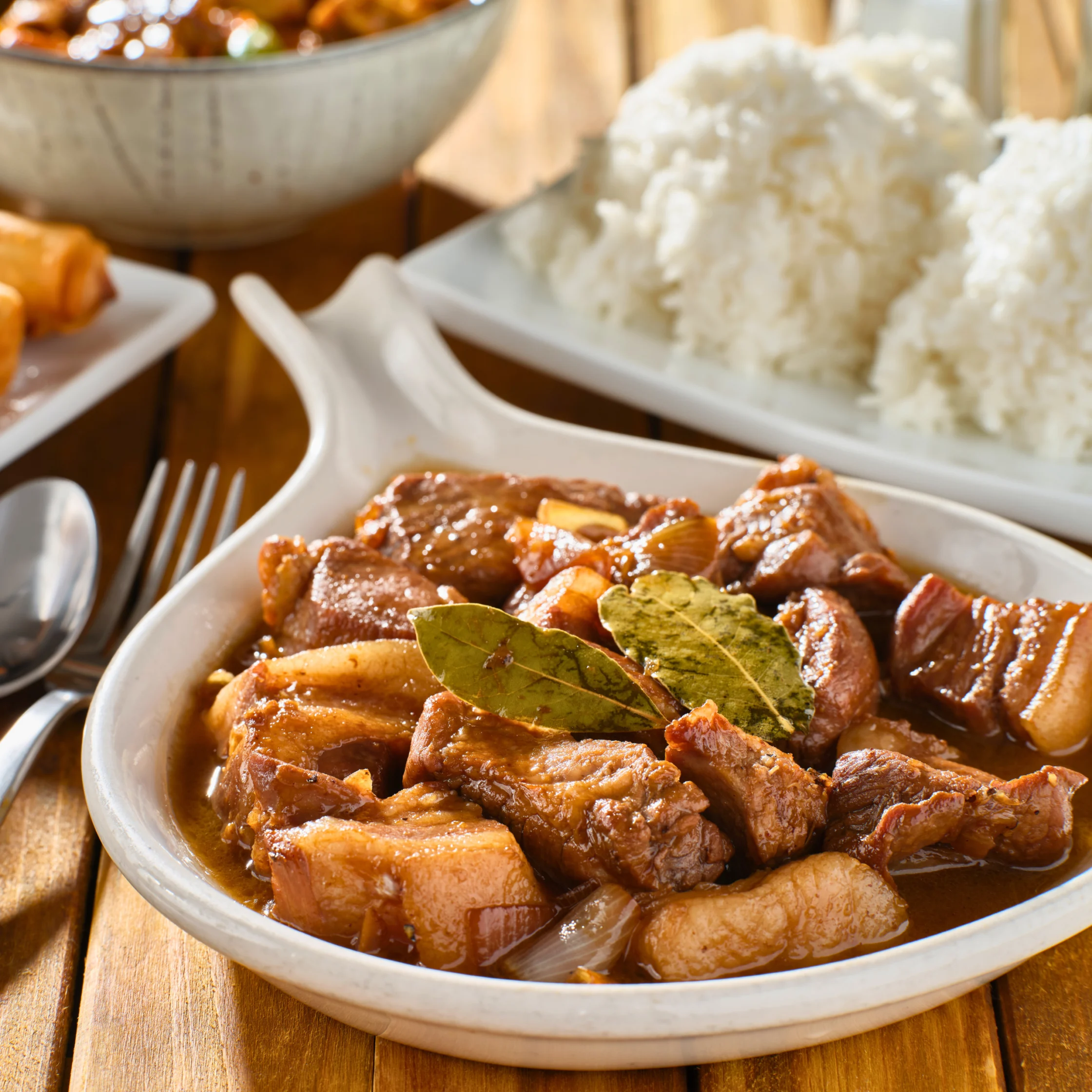 Bowl of Filipino pork adobo in a rich brown sauce, garnished with bay leaves and served with white rice.