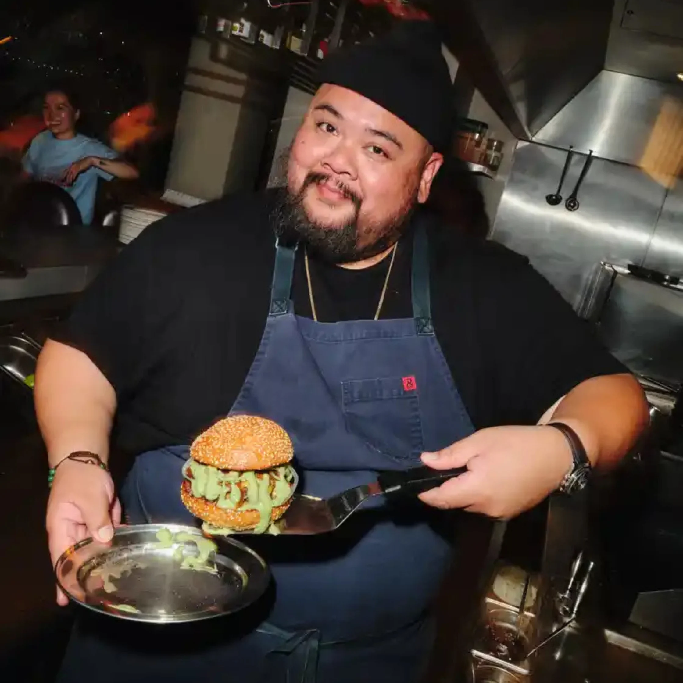 Alvin Cailan stands in a restaurant kitchen wearing a dark apron and cap while presenting a burger balanced on a spatula with green sauce dripping from the sides.