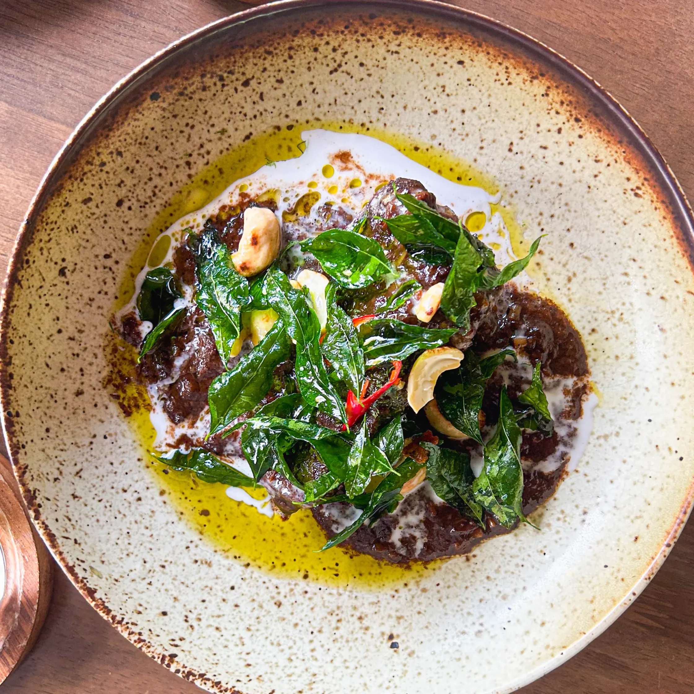 Slow-braised Angus beef cheek in a rich chocolate-massaman curry sauce, topped with crispy curry leaves, roasted cashews, and red chili, served in a shallow ceramic bowl.