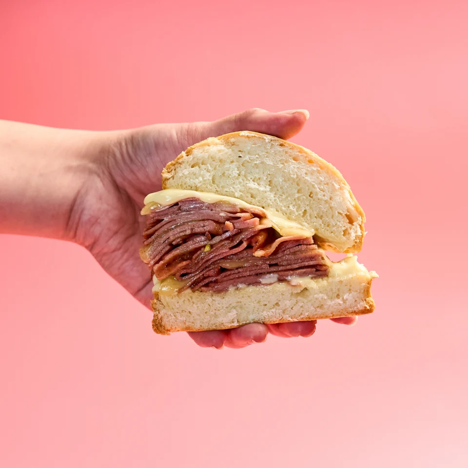 Anthony Bourdain-inspired mortadella sandwich stacked high with folded mortadella slices, melted provolone cheese, and mayonnaise on a soft round bread roll, served on a light blue plate against a pink background.