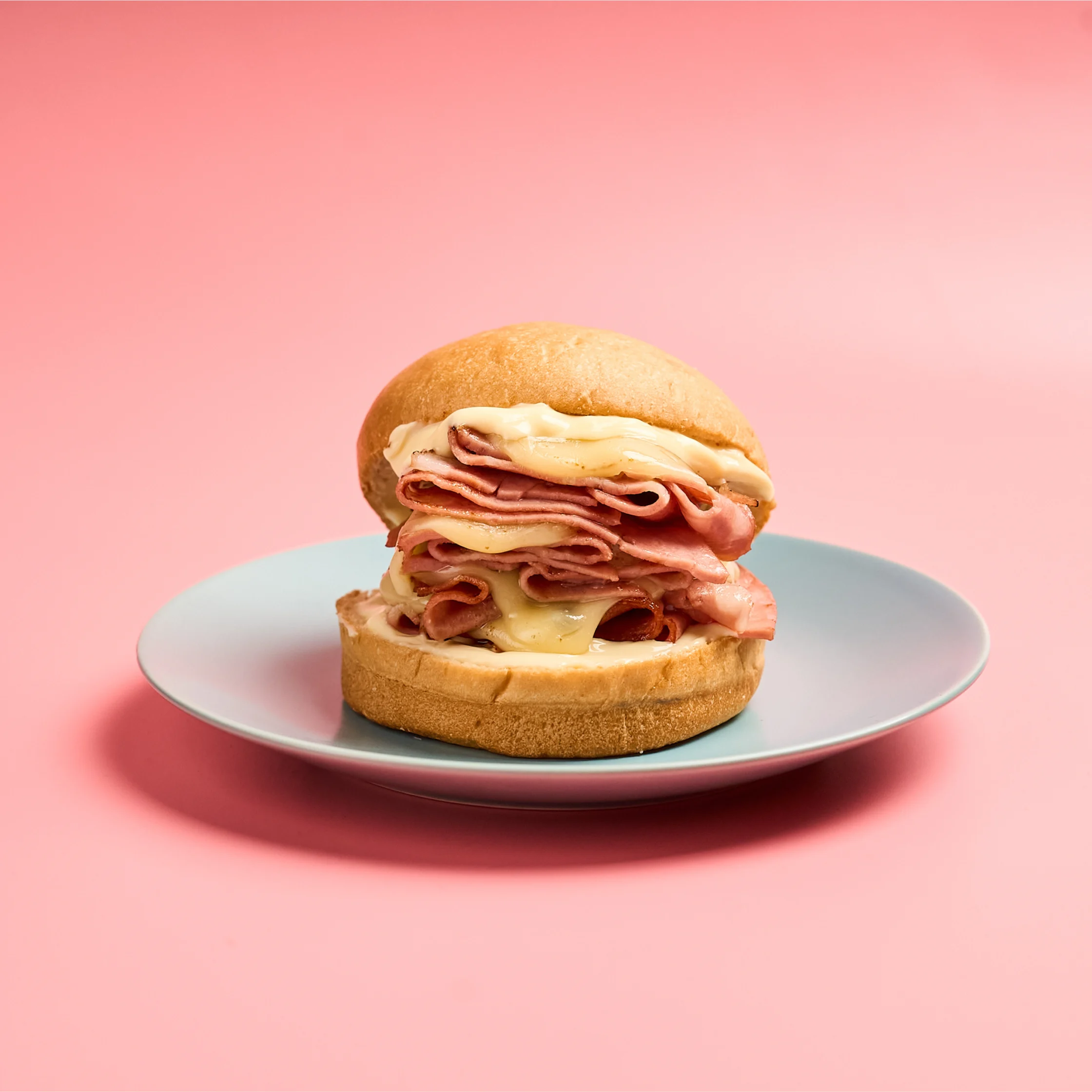 Anthony Bourdain-inspired mortadella sandwich stacked high with folded mortadella slices, melted provolone cheese, and mayonnaise on a soft round bread roll, served on a light blue plate against a pink background.