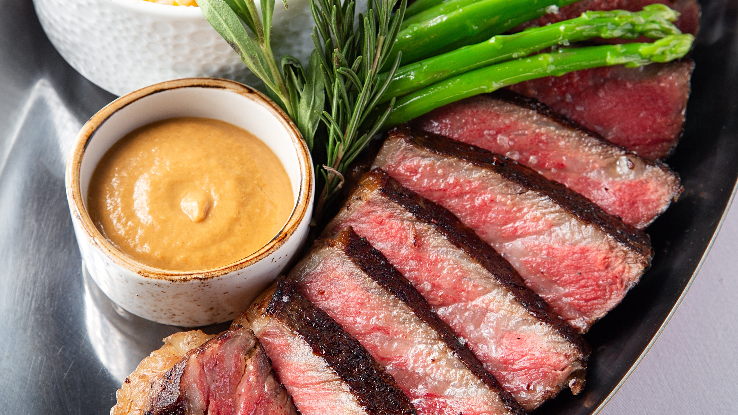 BRONTË bone-in New York strip steak sliced medium-rare and served with asparagus, rosemary, a creamy sauce, and a side of corn and brie gratin.