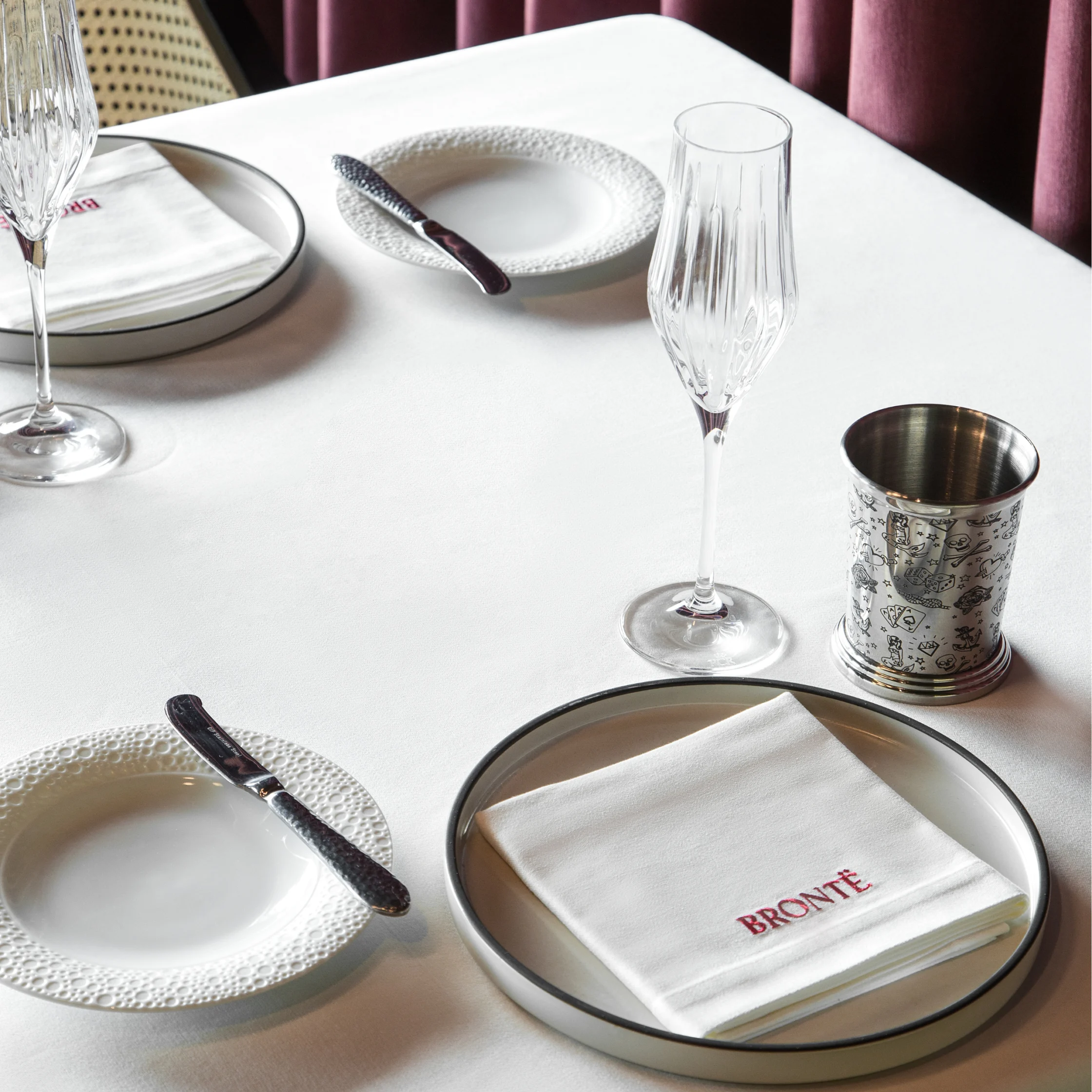 Detailed table setting at BRONTË featuring textured white plates, crystal stemware, silver-toned cups, cutlery, and a folded napkin embroidered with the BRONTË name.
