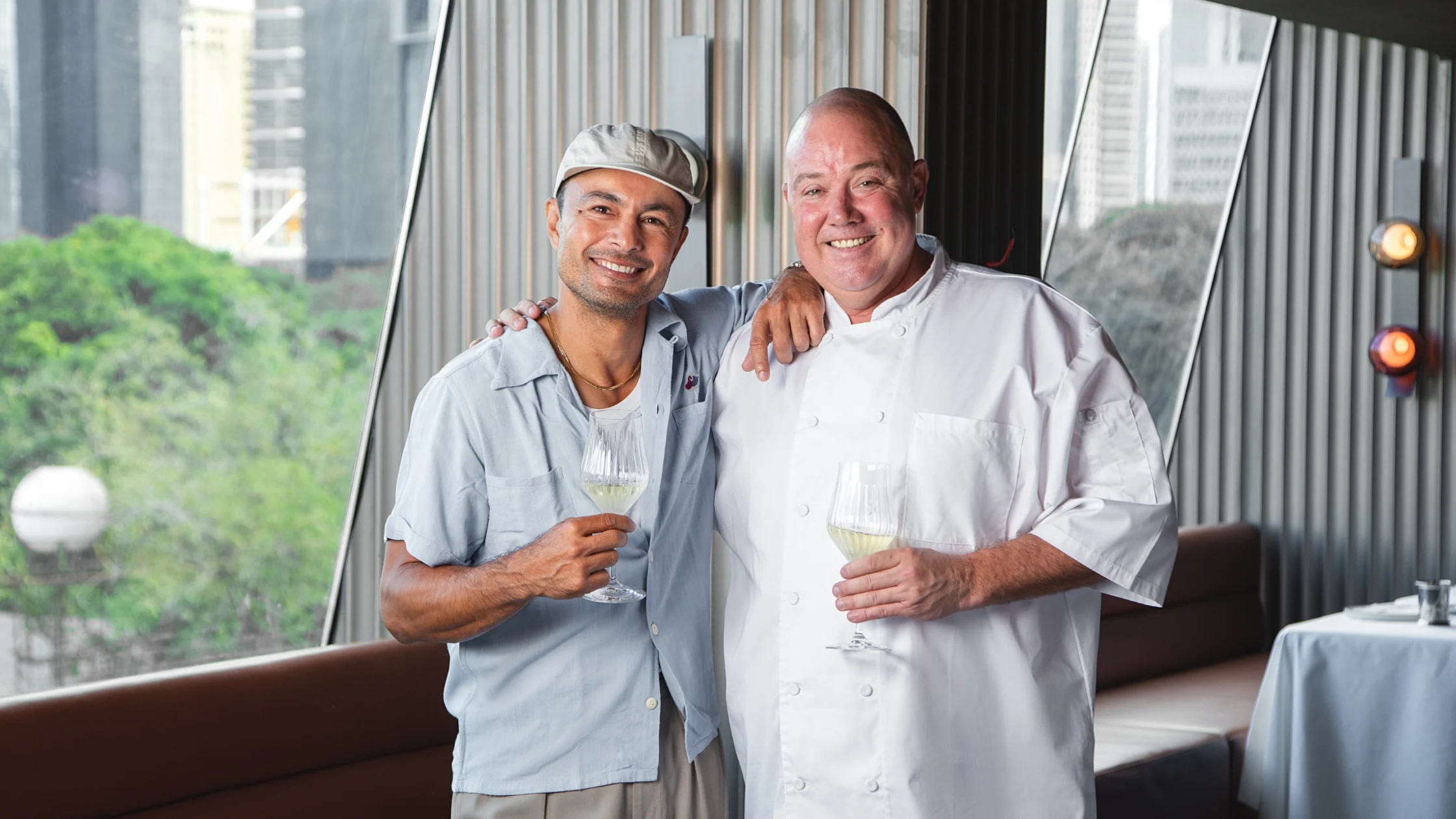Owners Derek Ramsay & Wade Watson smiling and holding glasses of white wine inside BRONTË, standing near large windows with views of greenery and the city skyline.