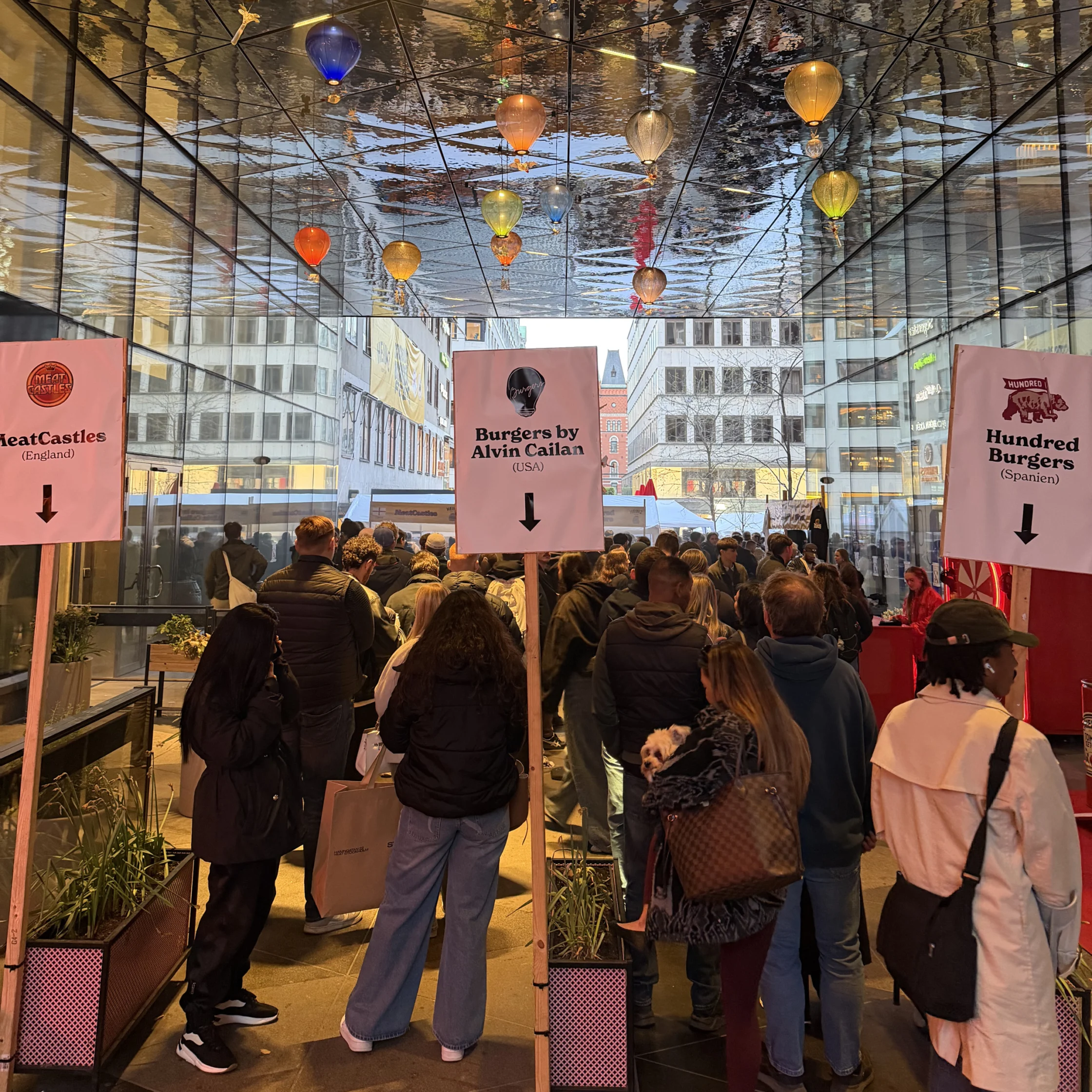 A crowd gathers beneath reflective ceilings and colorful hanging lights at a burger event entrance with signage pointing to “Burgers by Alvin Cailan"