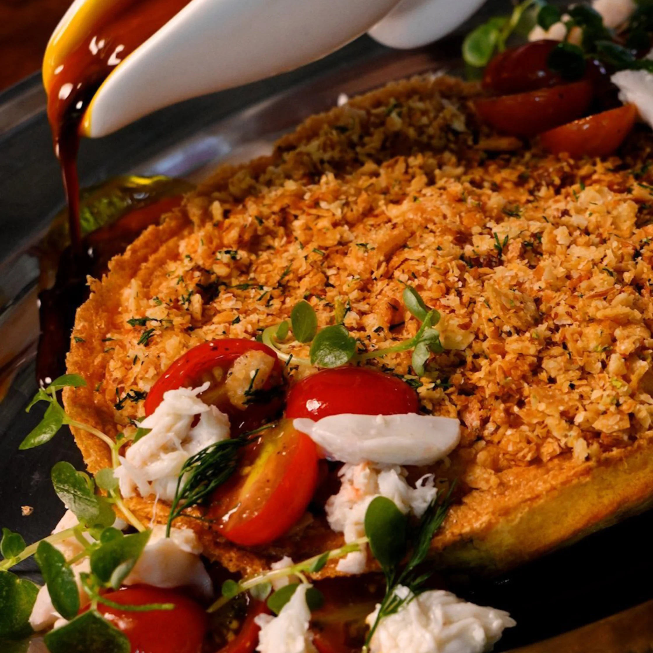 Close-up of a Filipino savory tart or pie with a crisp golden crust, topped with toasted crumbs, cherry tomatoes, herbs, and pieces of seafood, while a dark sauce is being poured over the side.