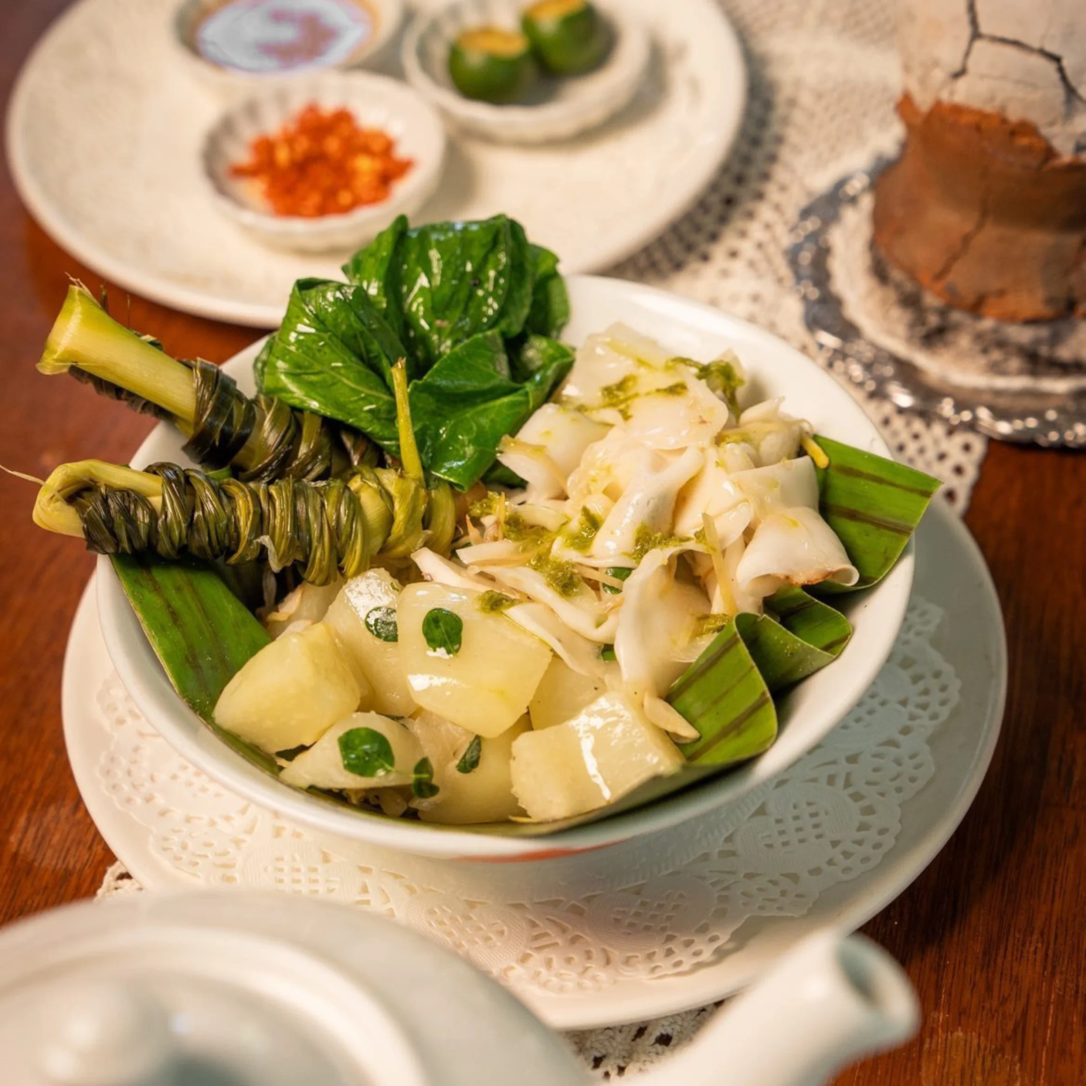 A bowl of Filipino-style vegetable and seafood dish served on banana leaves, featuring leafy greens, squid, and chunks of root vegetables, with wrapped pandan leaf bundles on top and calamansi with chopped chilies in the background.