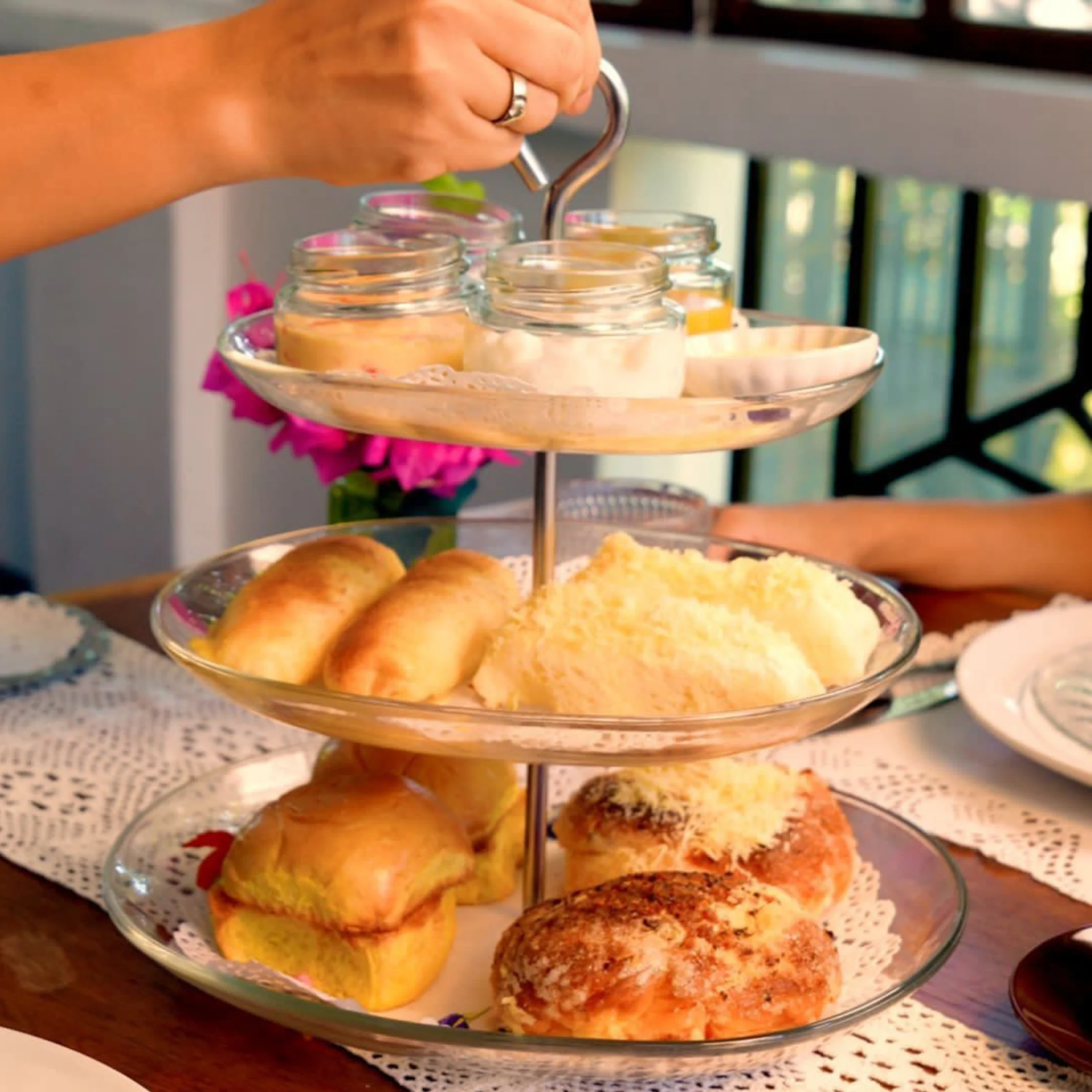 A three-tier stand of Filipino merienda pastries and breads, including ensaymada topped with grated cheese, soft bread rolls, and small jars of sweet spreads.